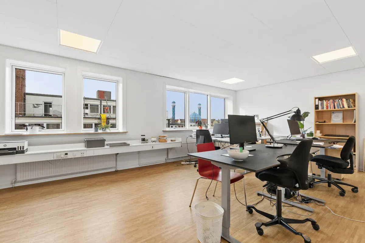 Kontor med moderne interiør, skriveborde, stolpe og vinduer med udsigt til bygninger og tårne i baggrunden