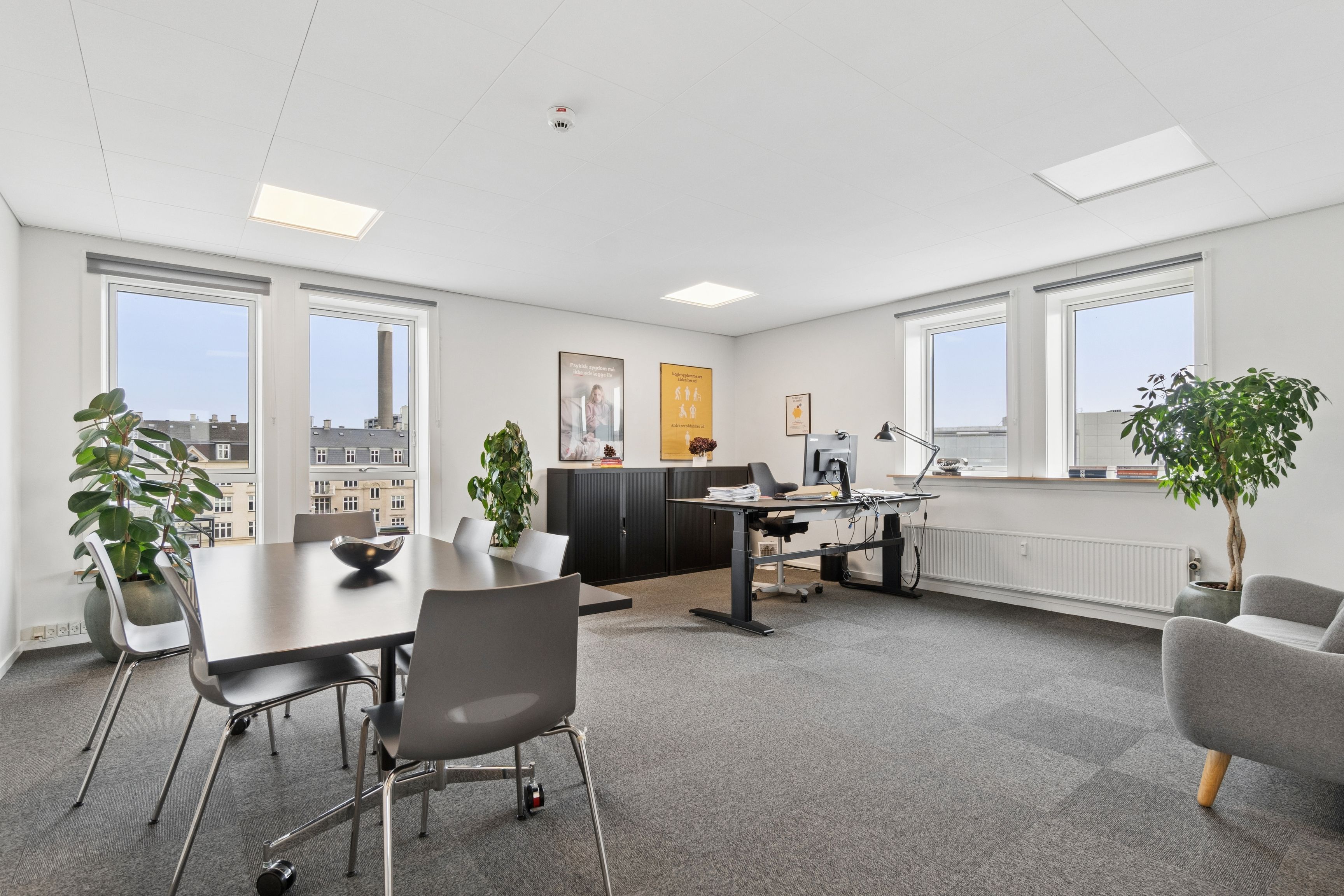 Kontorrum med store vinduer og højt loft, møbler og plante. To skriveborde med computer og lampe samt stol. Bord med skål og flere stole.