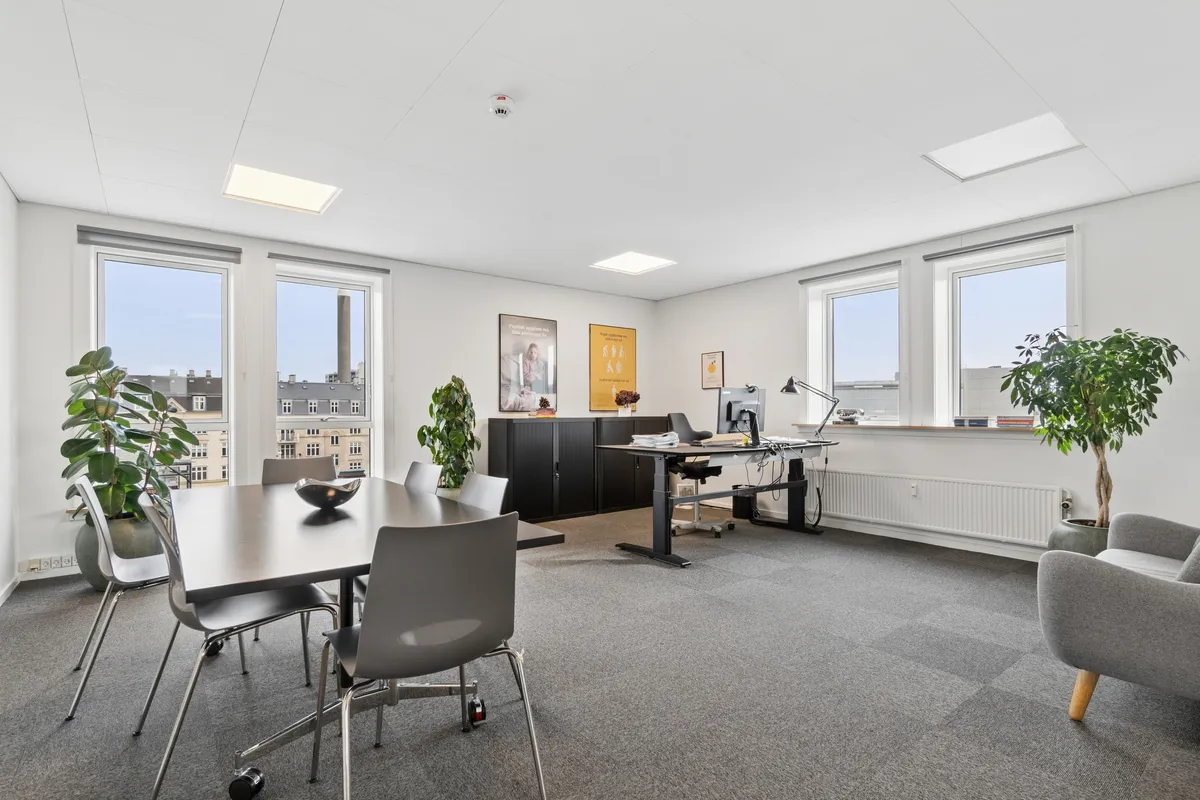 Kontorrum med store vinduer og højt loft, møbler og plante. To skriveborde med computer og lampe samt stol. Bord med skål og flere stole.