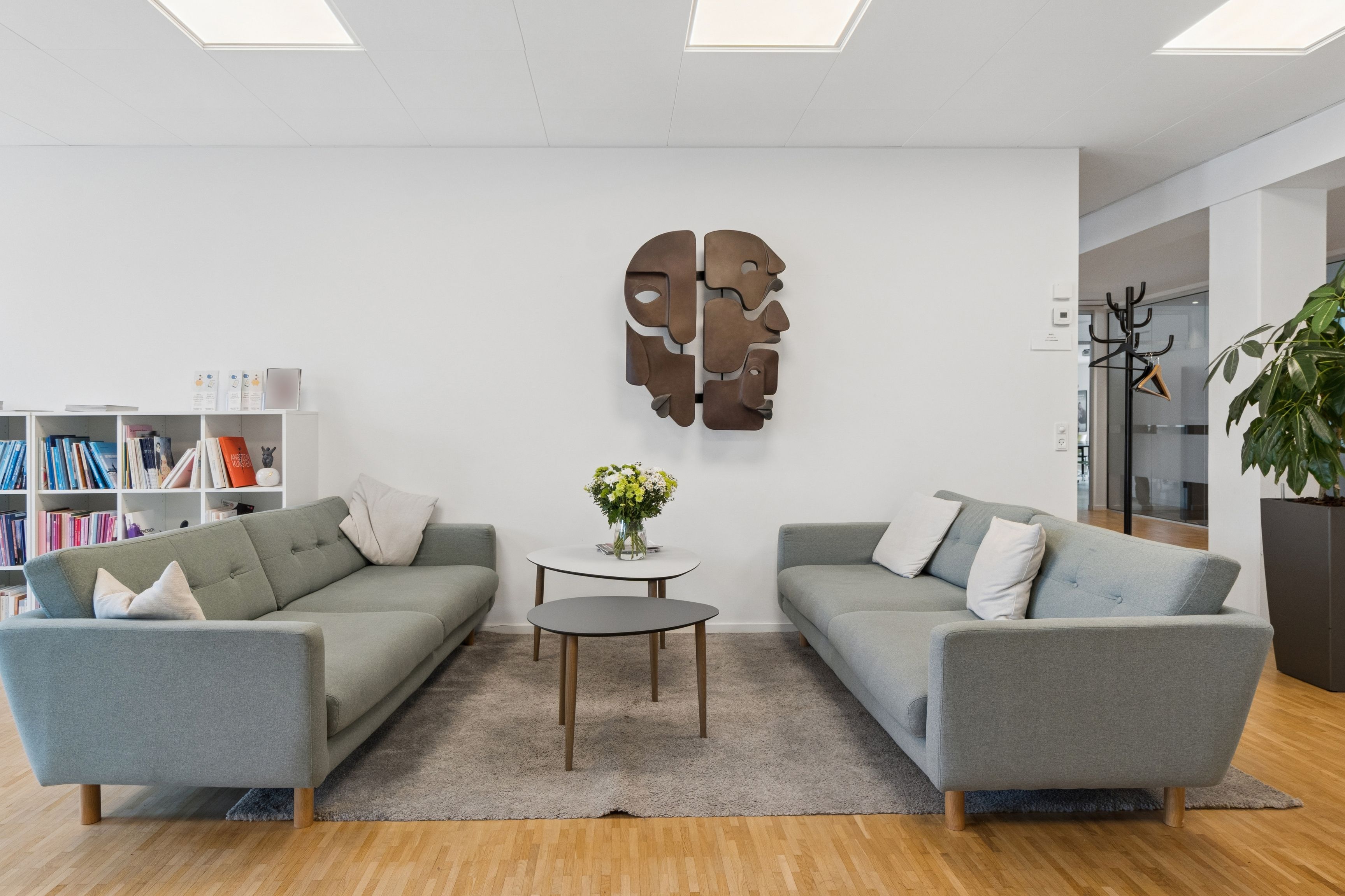 To sovesofaer med pudesæt og to bord med blomster i en stue med en stor plante og et stort vægmaleri med menneskelige ansigter