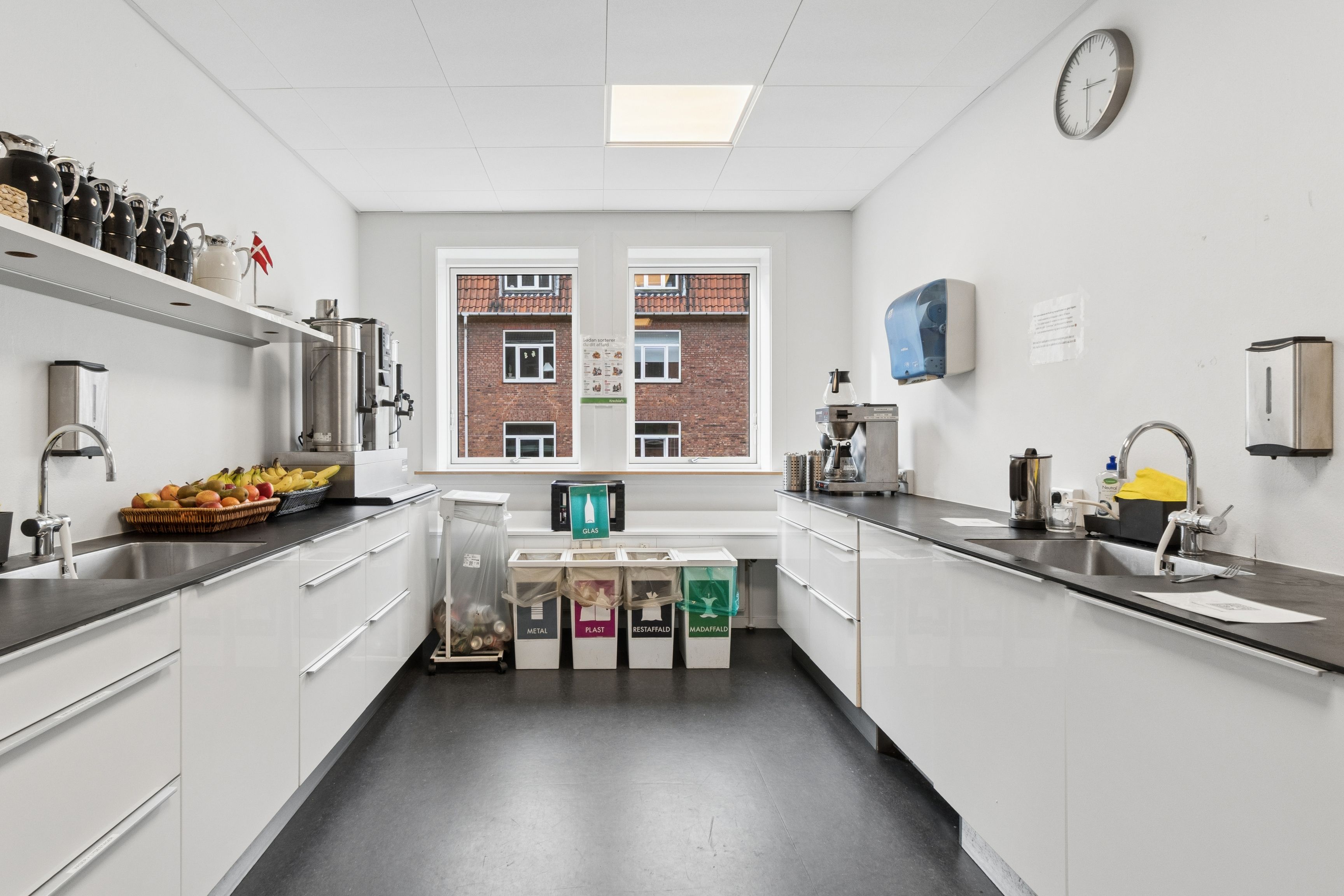 Indkøbskøkken med lyse krydderier, frugtkurv, kaffemaskine og flere skraldspande for forskellige materialer på en hvid belægningsgulv