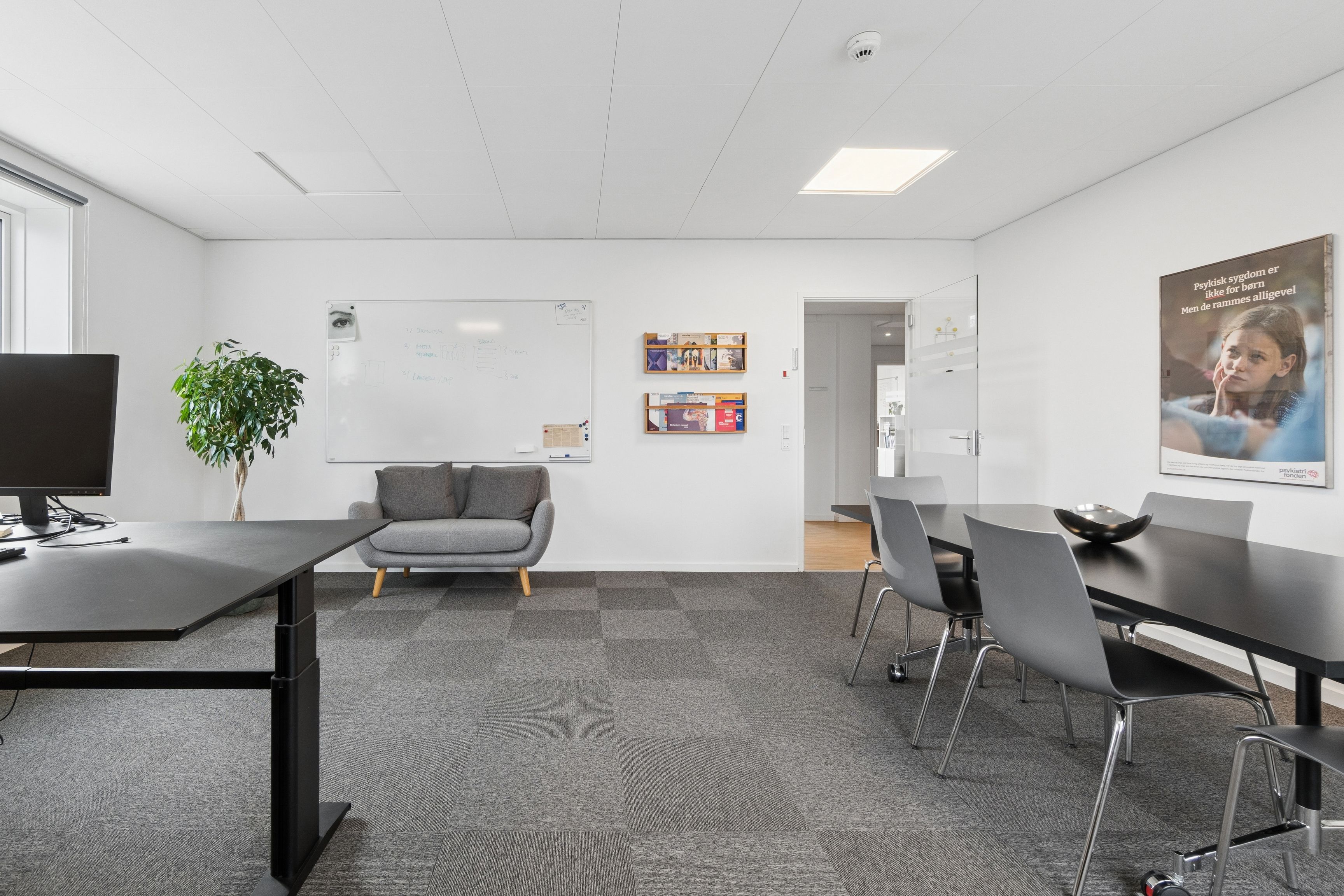 Et enkelt kontorrum med højtloft og møbler i sort og gråt, samt en sofa og en plante. På væggen er en whiteboard og et plakat med en pige.