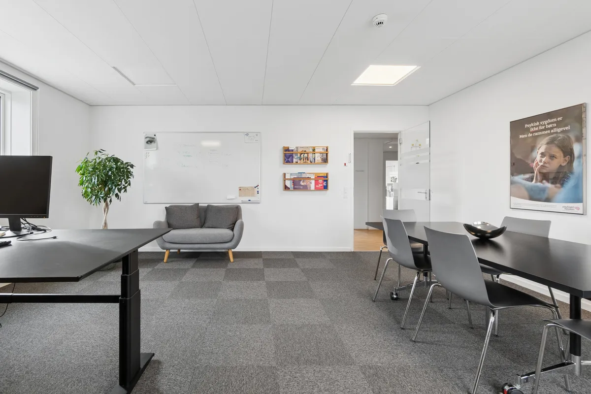 Et enkelt kontorrum med højtloft og møbler i sort og gråt, samt en sofa og en plante. På væggen er en whiteboard og et plakat med en pige.