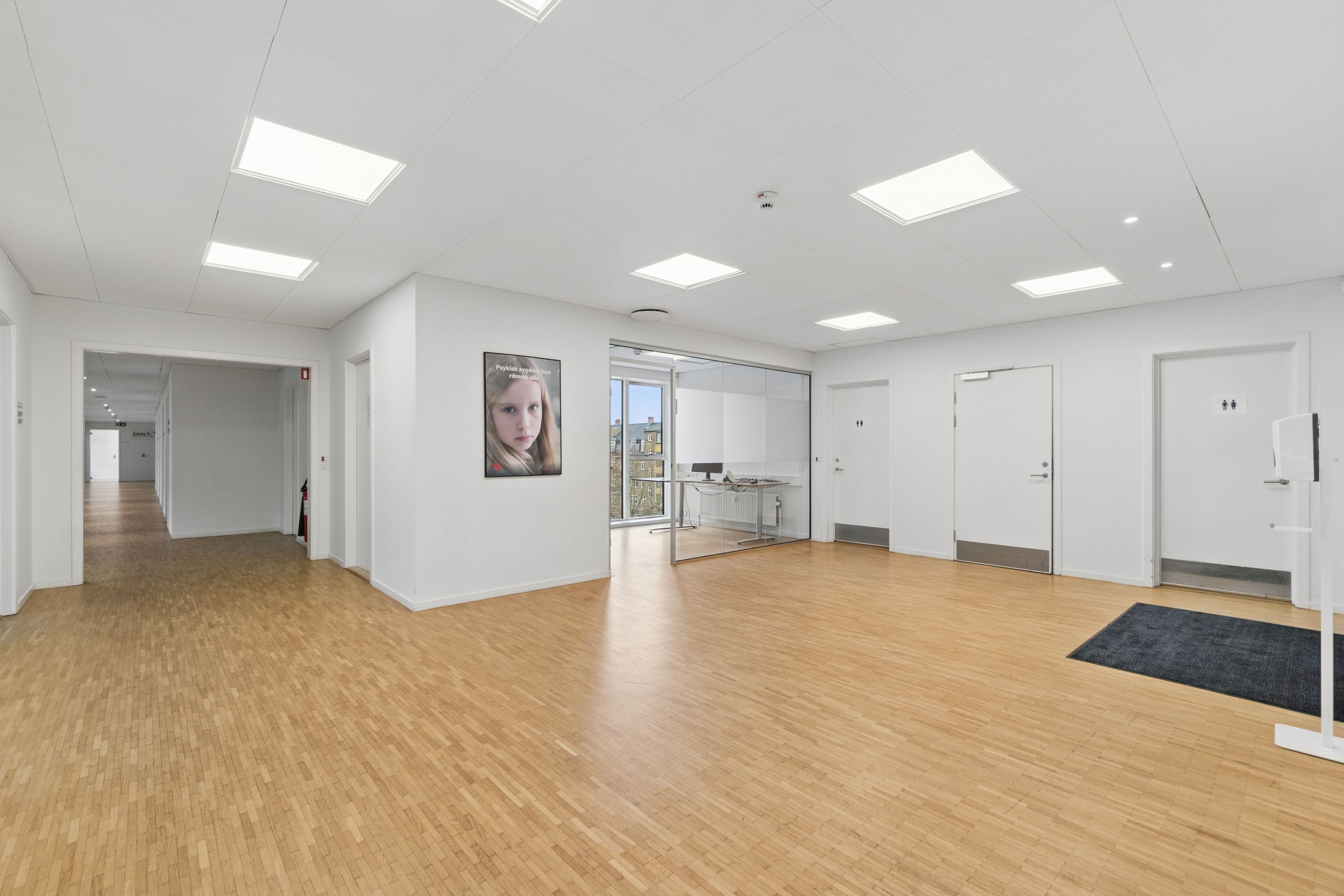 Et lyse kontor med hvide vægge og loft. Der er et gulv med træpaneler. Der er en dør med et toiletskilt og en udgang til en veranda med et bord og en stol.