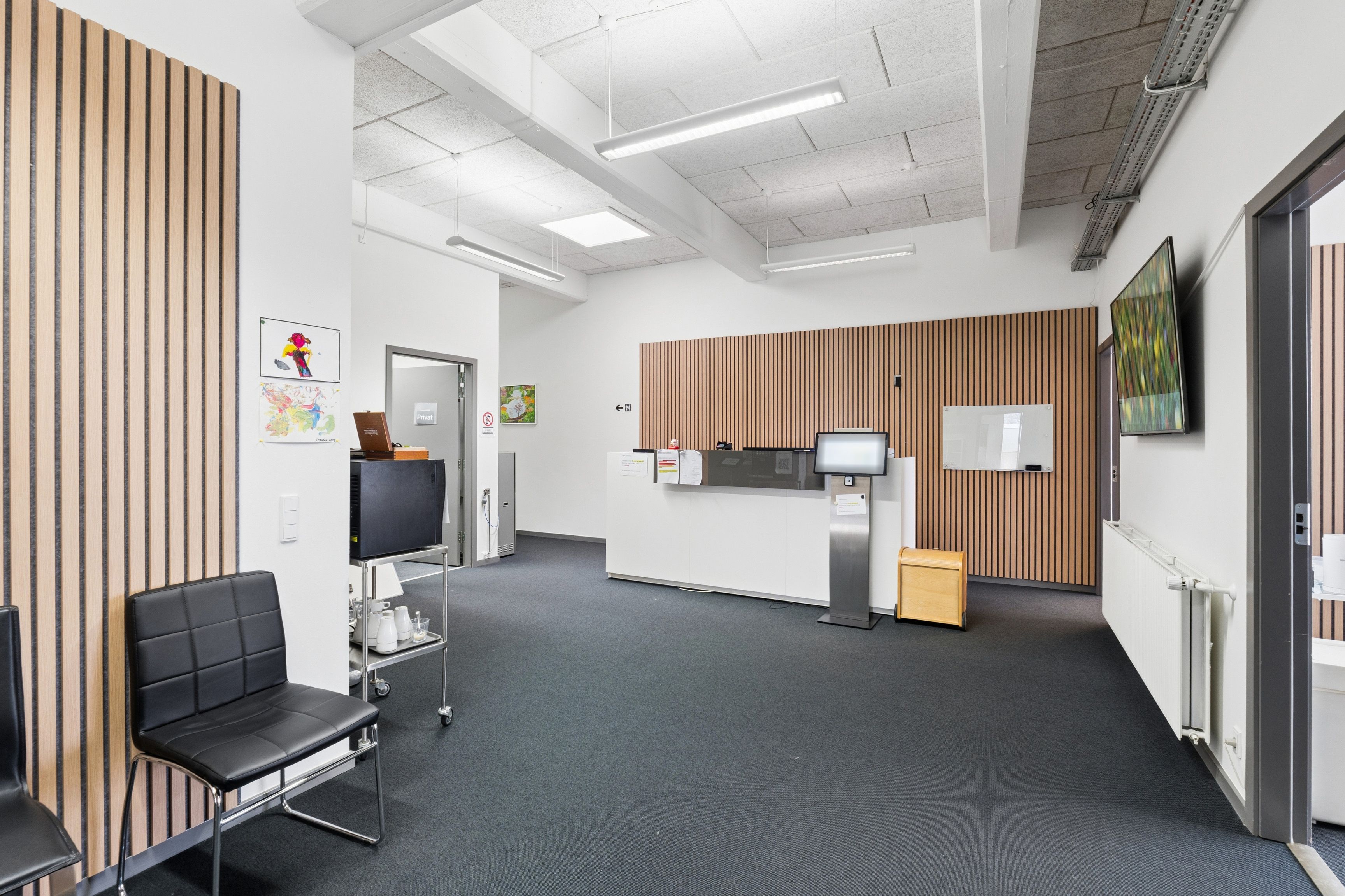 Lyst kontor med højt til loftet og moderne møbler. Foran er læderstole, træskabe og en ståbord med monitor. Der er hængende lysekroner og flere malerier på væggen