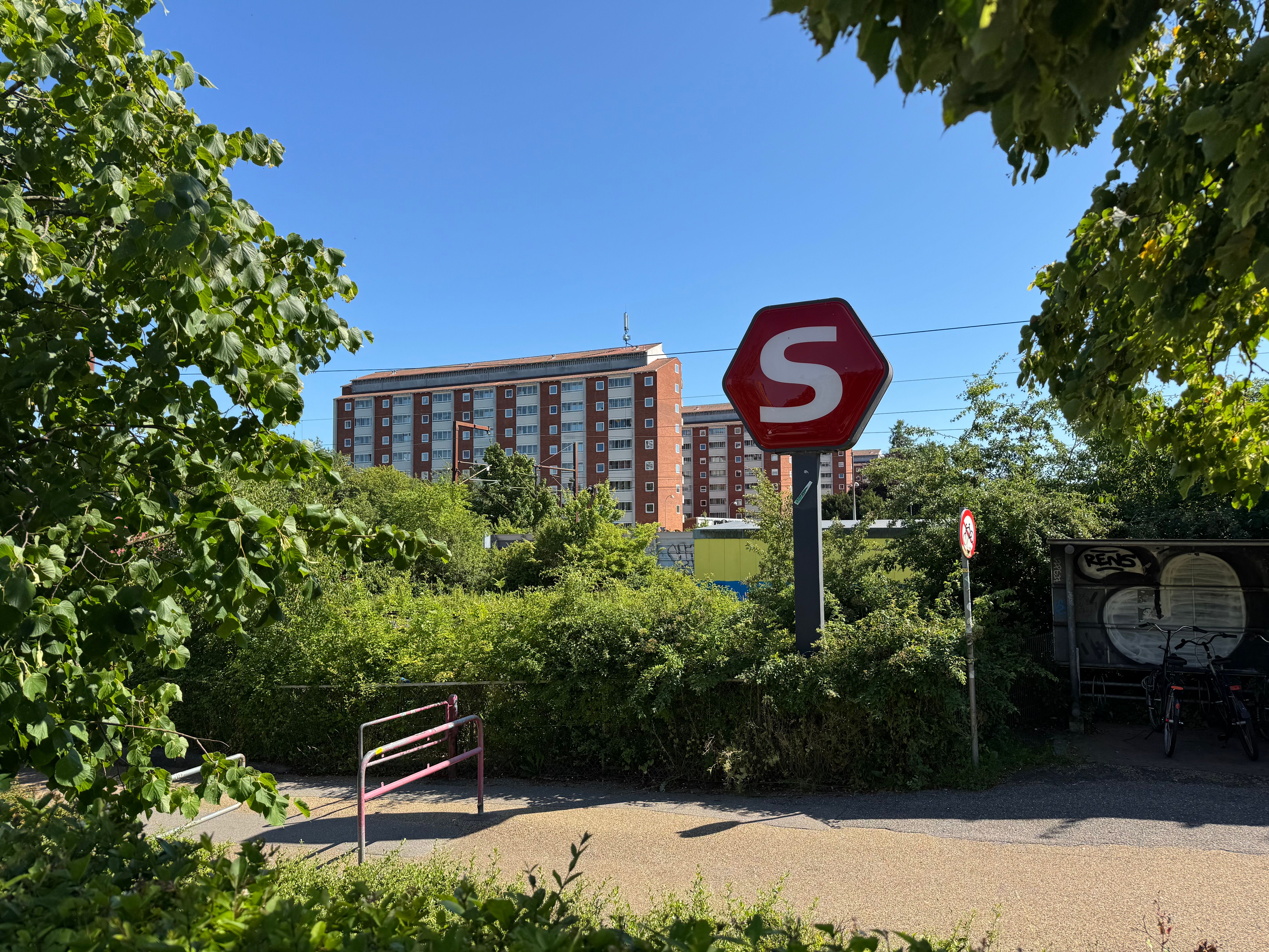 En stor bygning med mange vinduer og en stor stopskilt med bogstavet S i midten, omgivet af buske og træer. Bag bygningen er der to parkerede cykler.