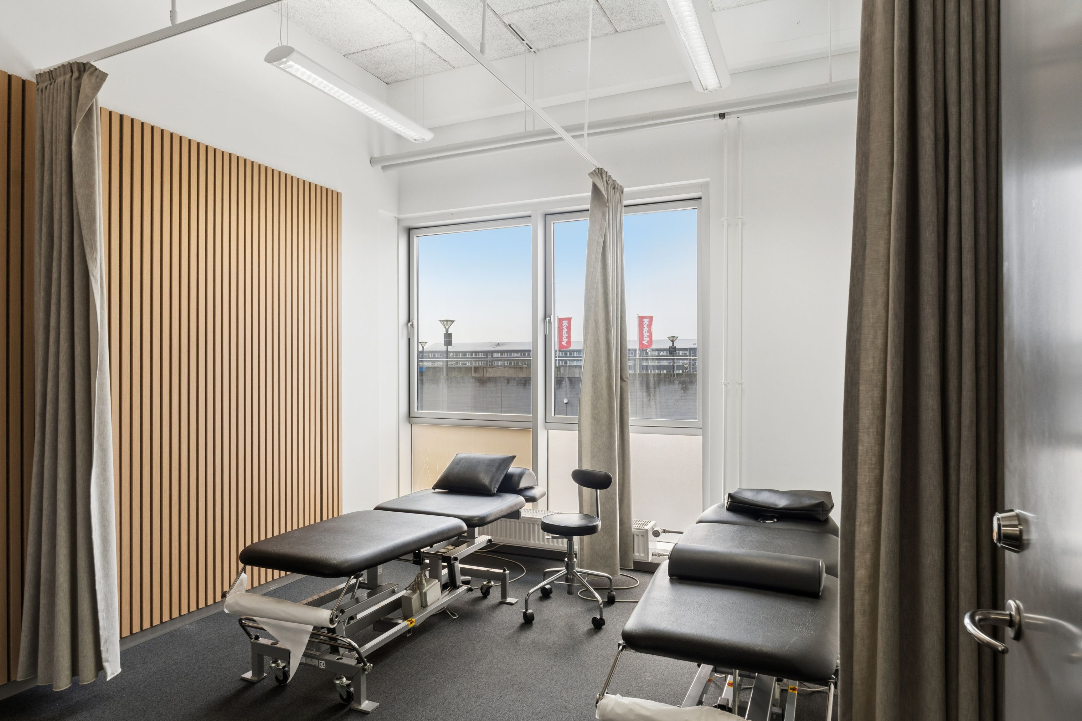 Lyst behandlerværelse med højt til loftet og smukke træpaneler i en moderne kontorbygning med udsigt til vandet og byen i baggrunden