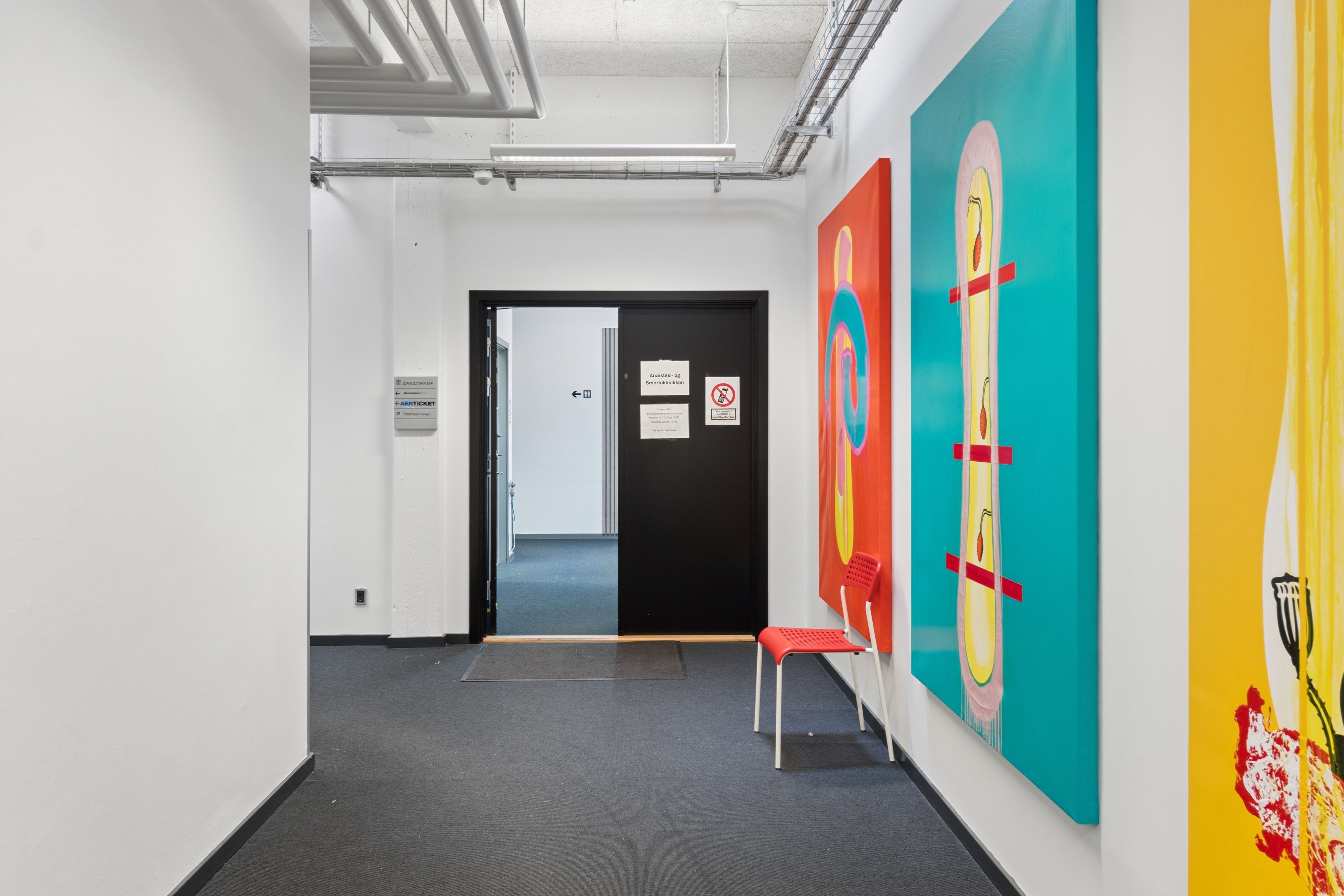 En tom, lyse gang med lysegule vægge, hvor der er to store malerier og en rød stol. Der er to døre og en væg med skilte