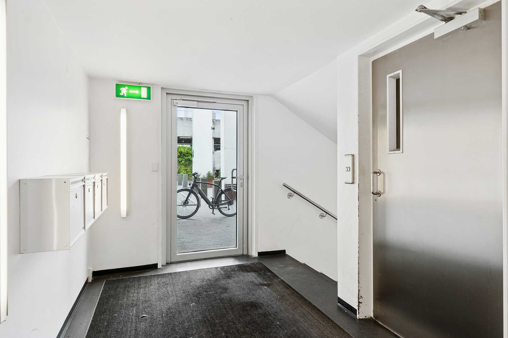 Modernt indgangsområde med hvidt vægpanel og glasdør, der giver udsigt til gården med cykler. Der er skriftligt og lysmæssigt brandsikringstilbehør og håndtag på den åbne dør.