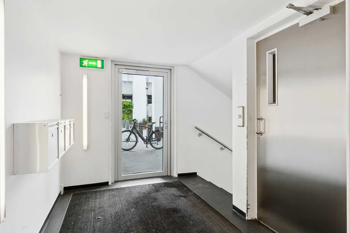 Modernt indgangsområde med hvidt vægpanel og glasdør, der giver udsigt til gården med cykler. Der er skriftligt og lysmæssigt brandsikringstilbehør og håndtag på den åbne dør.