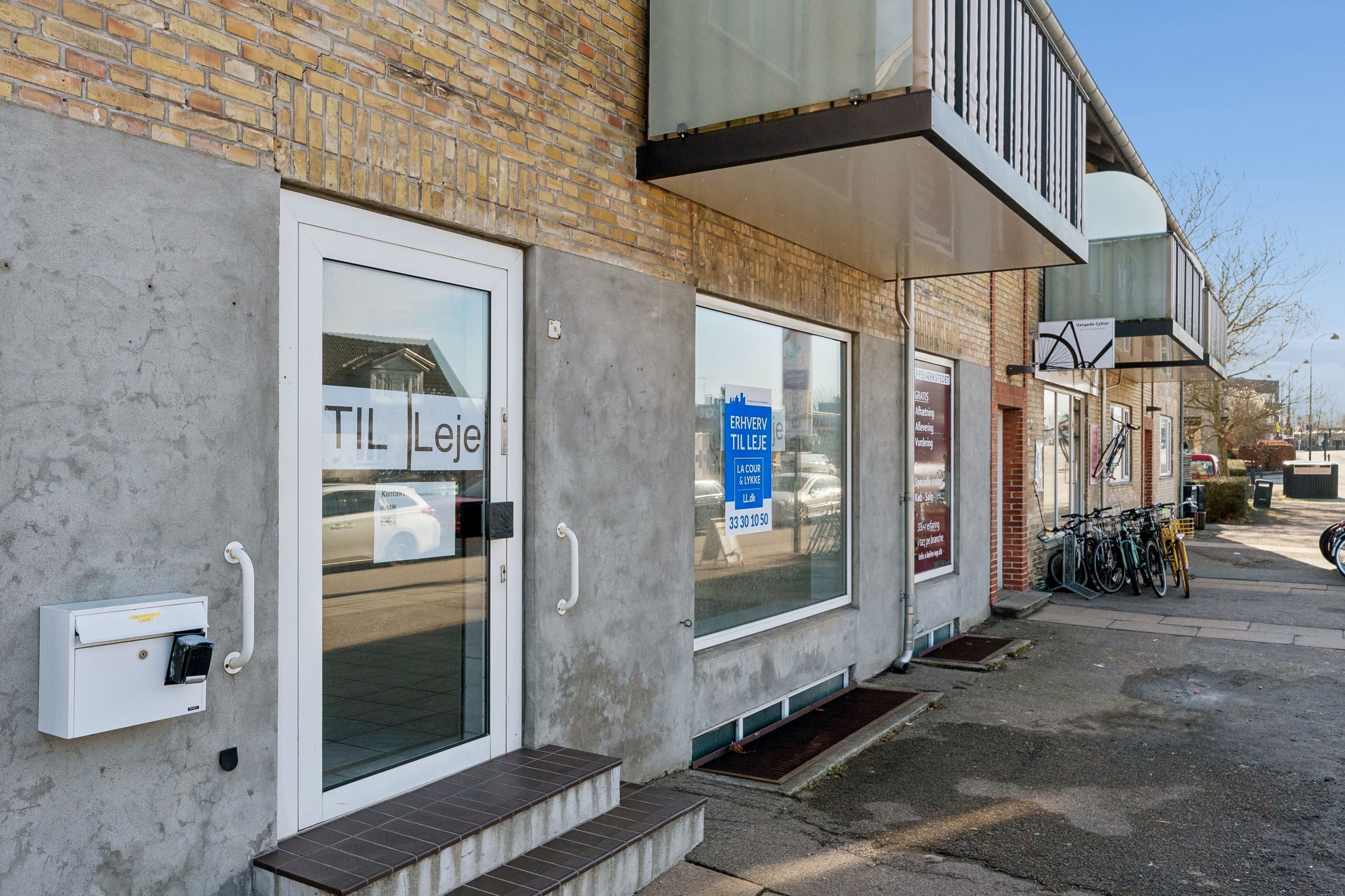 Butikstil leje på Vangedevej 131 i Gentofte. Vindue med tekst "Til leje". Parkerede cykler og biler i baggrunden. Lys og klar himmel.