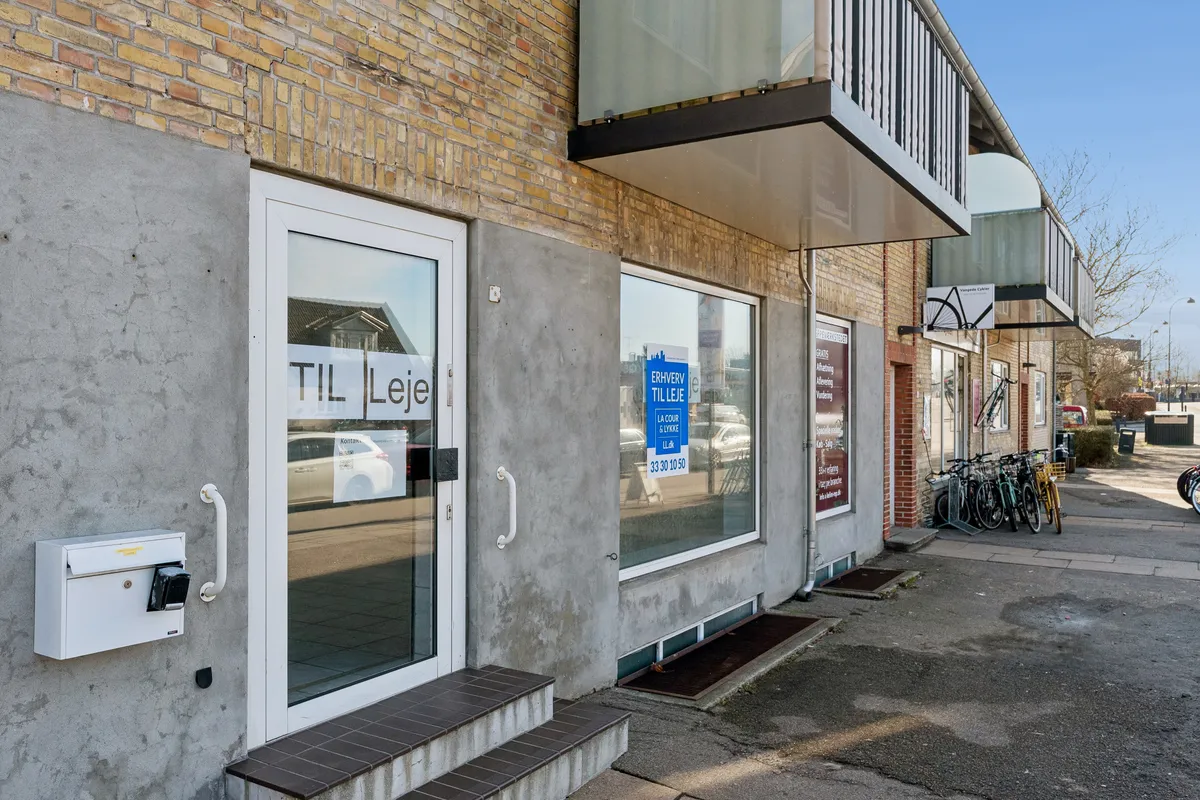 Butikstil leje på Vangedevej 131 i Gentofte. Vindue med tekst "Til leje". Parkerede cykler og biler i baggrunden. Lys og klar himmel.