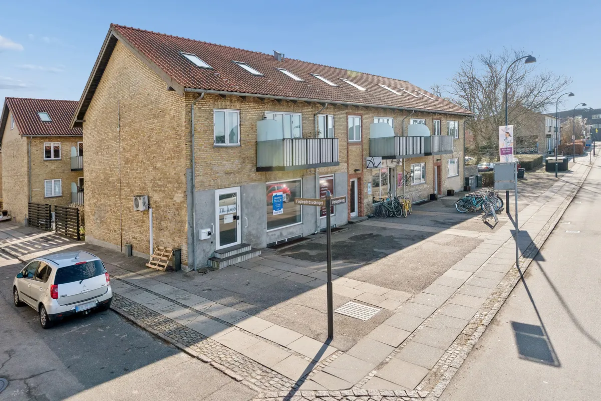 En højtliggende lejlighed til leje i en rød murstensbygning med store vinduer, udsigt til en parkeringsplads med parkerede cykler og biler samt en vej med gågade, fortov og parkerede biler