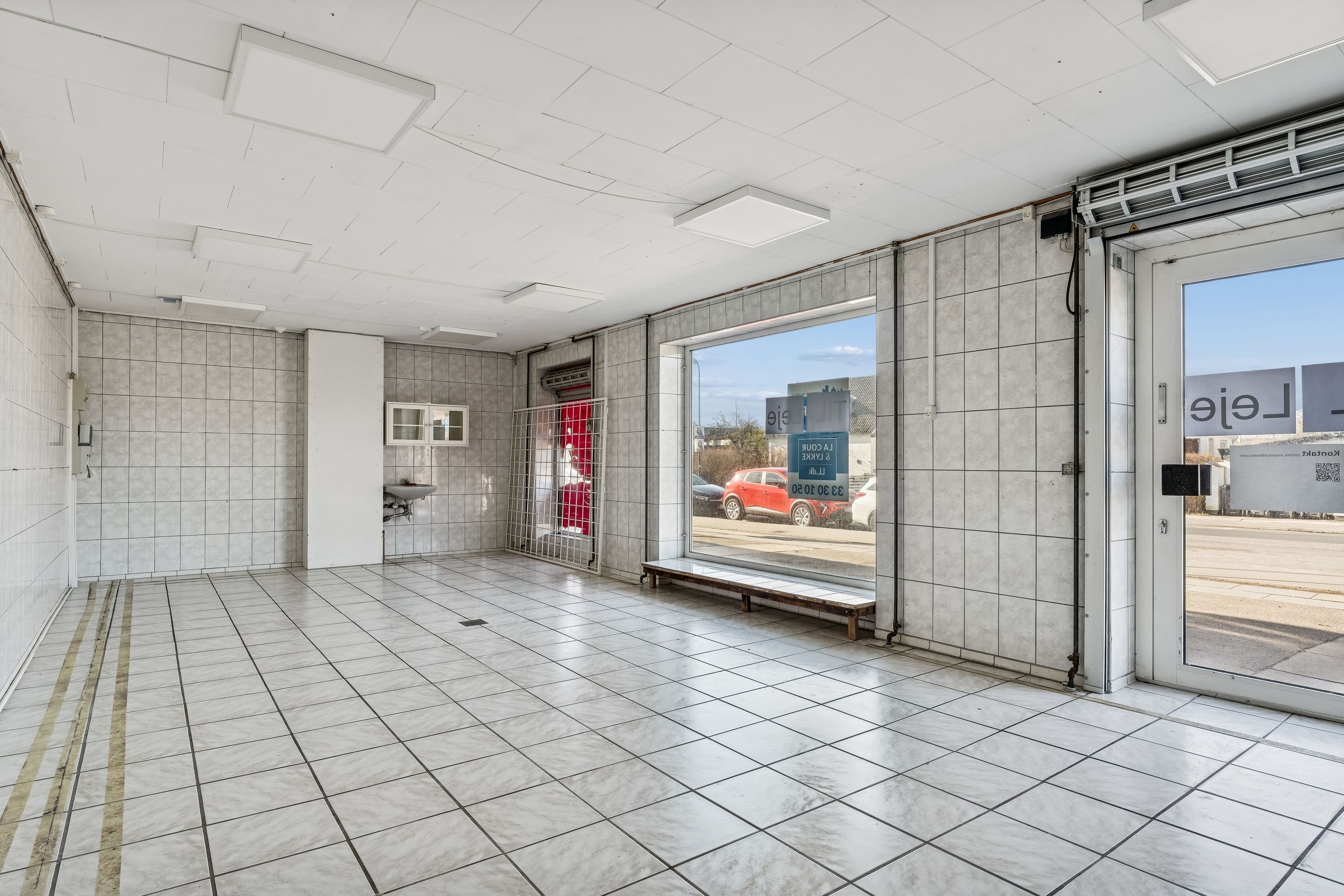 Ledig butikslokal med hvidt gulv og vægge, skur, en dør og et vindue med glas. Bag butikken er biler parkeret, og der er tekst på skiltene