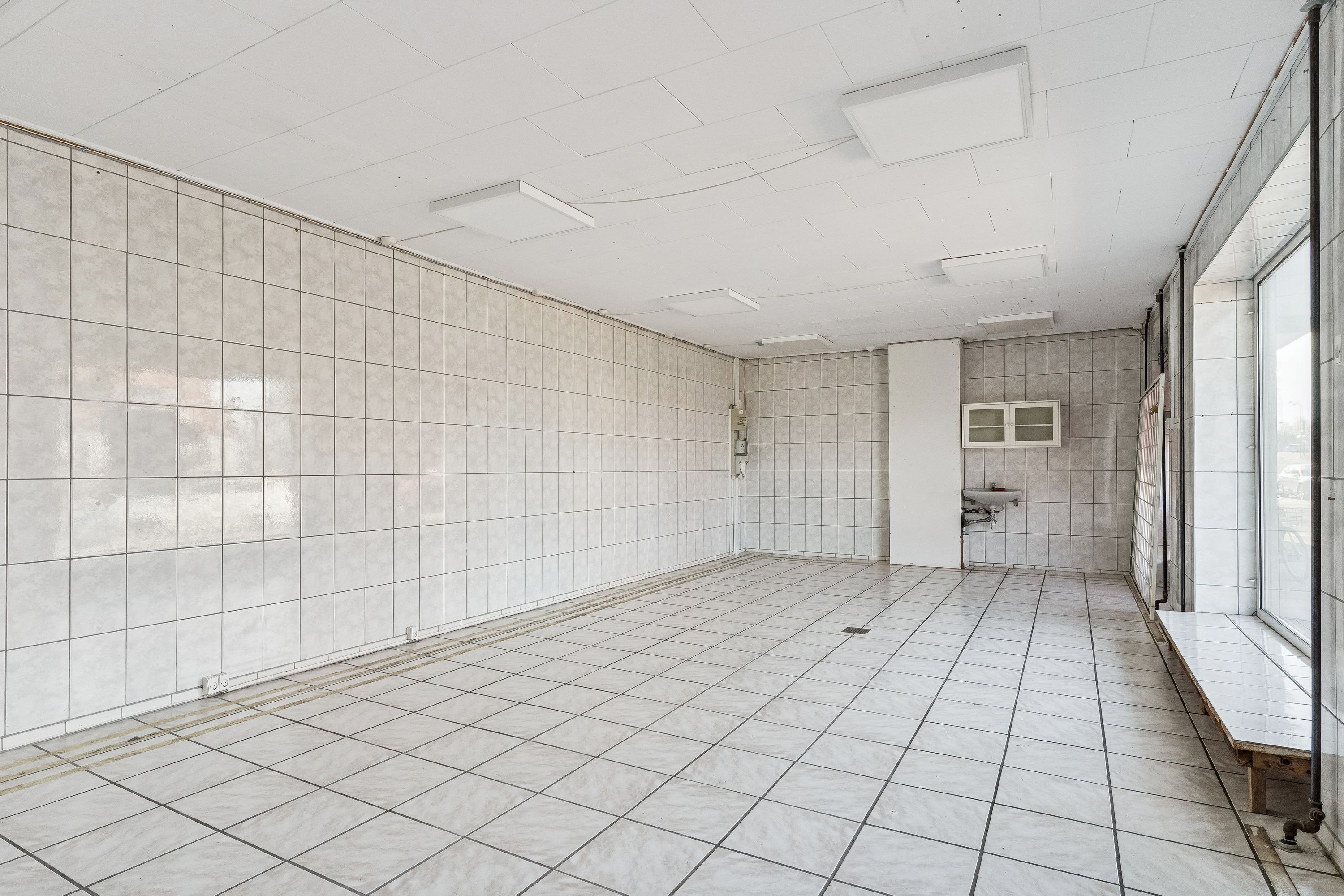 Ledig erhvervslokal med hvidt loft og gulv og flere vægge med flise. Der er også en kabinet og skab i hjørnet.