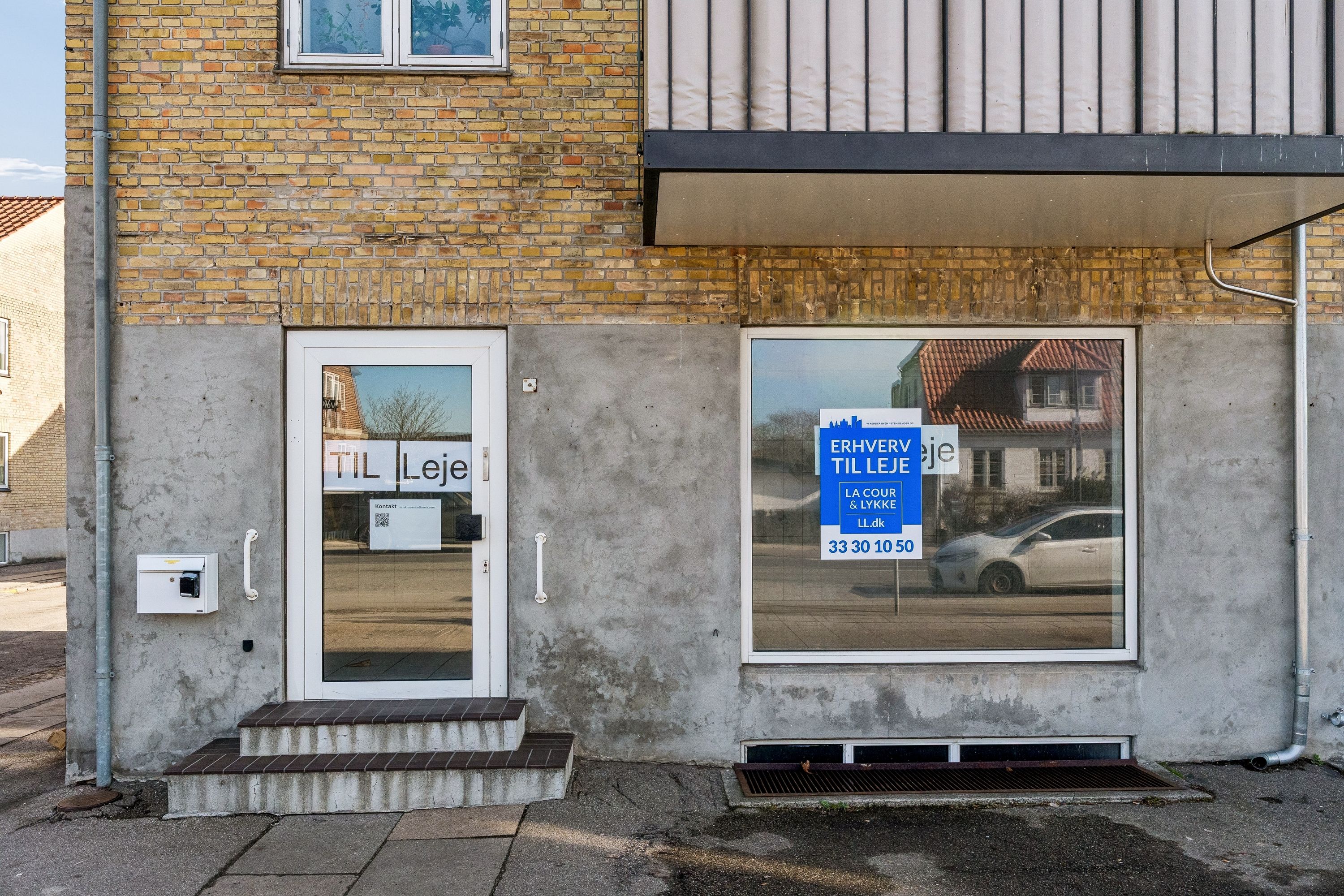 Bygning med to butiksruder og tekst på skiltet, hvor det står "Erhverv til leje" og "La Cour & Lykkeje" samt kontaktoplysninger og åbningstider