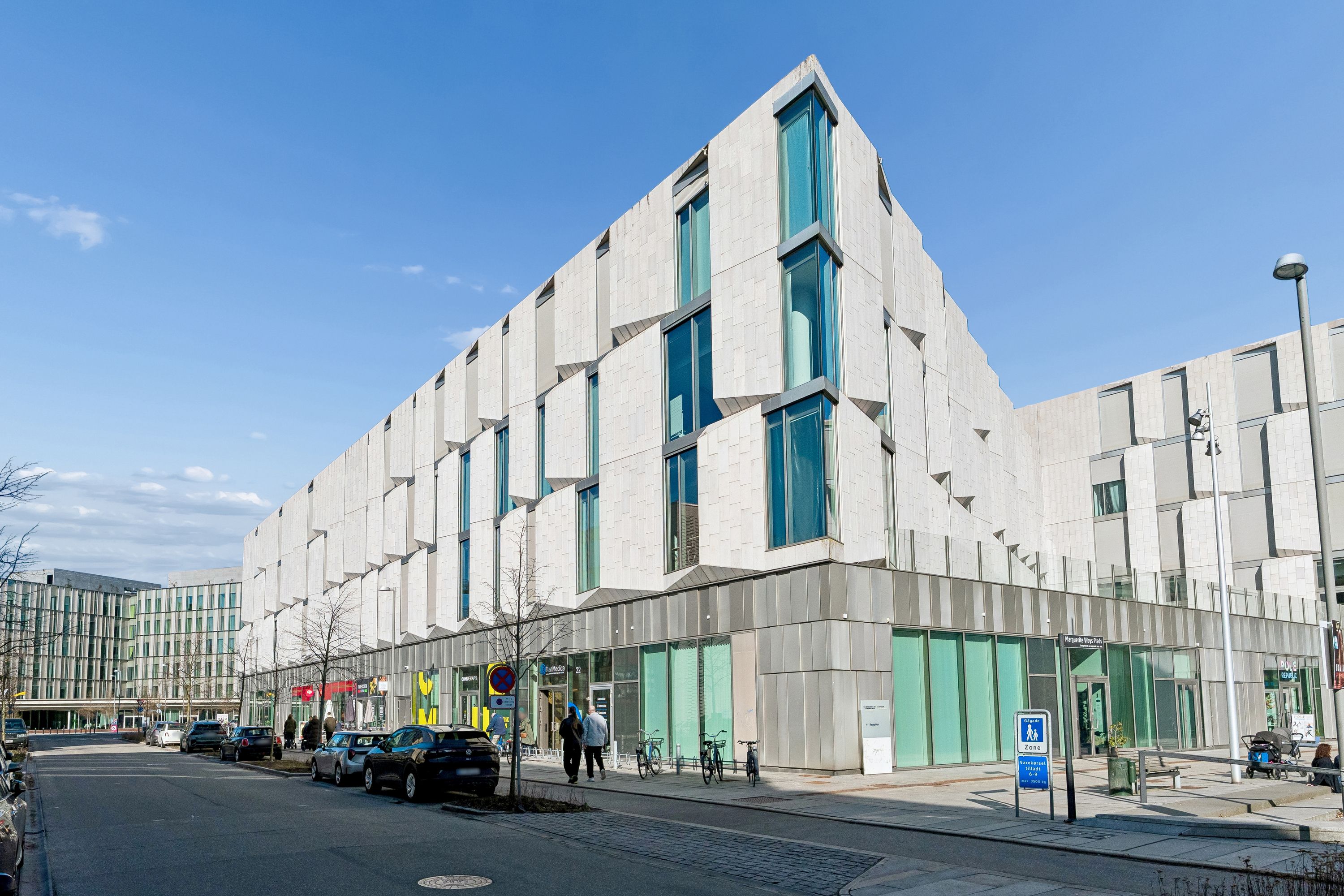 Flere personer gå forbi en stor hjørnebygning med moderne design og glasvinduer i Frederiksberg, hvor flere biler er parkeret langs gaden