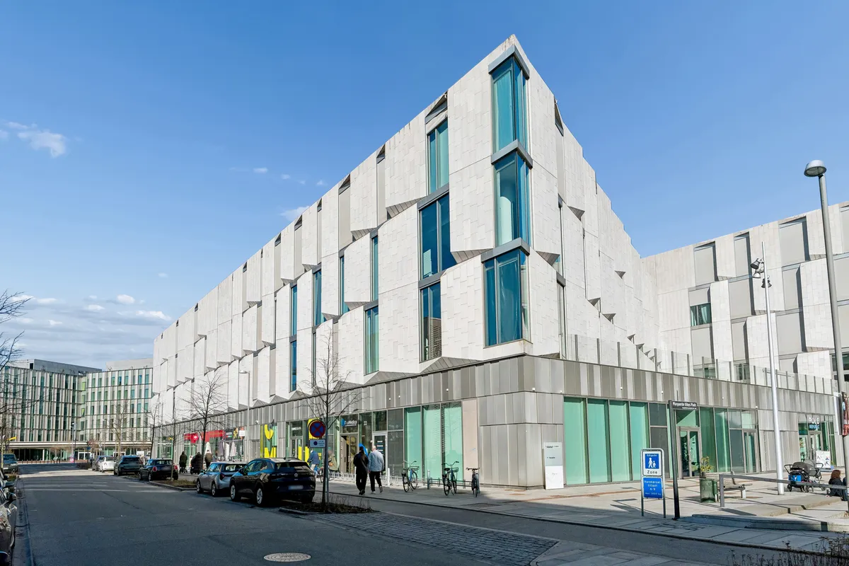 Flere personer gå forbi en stor hjørnebygning med moderne design og glasvinduer i Frederiksberg, hvor flere biler er parkeret langs gaden