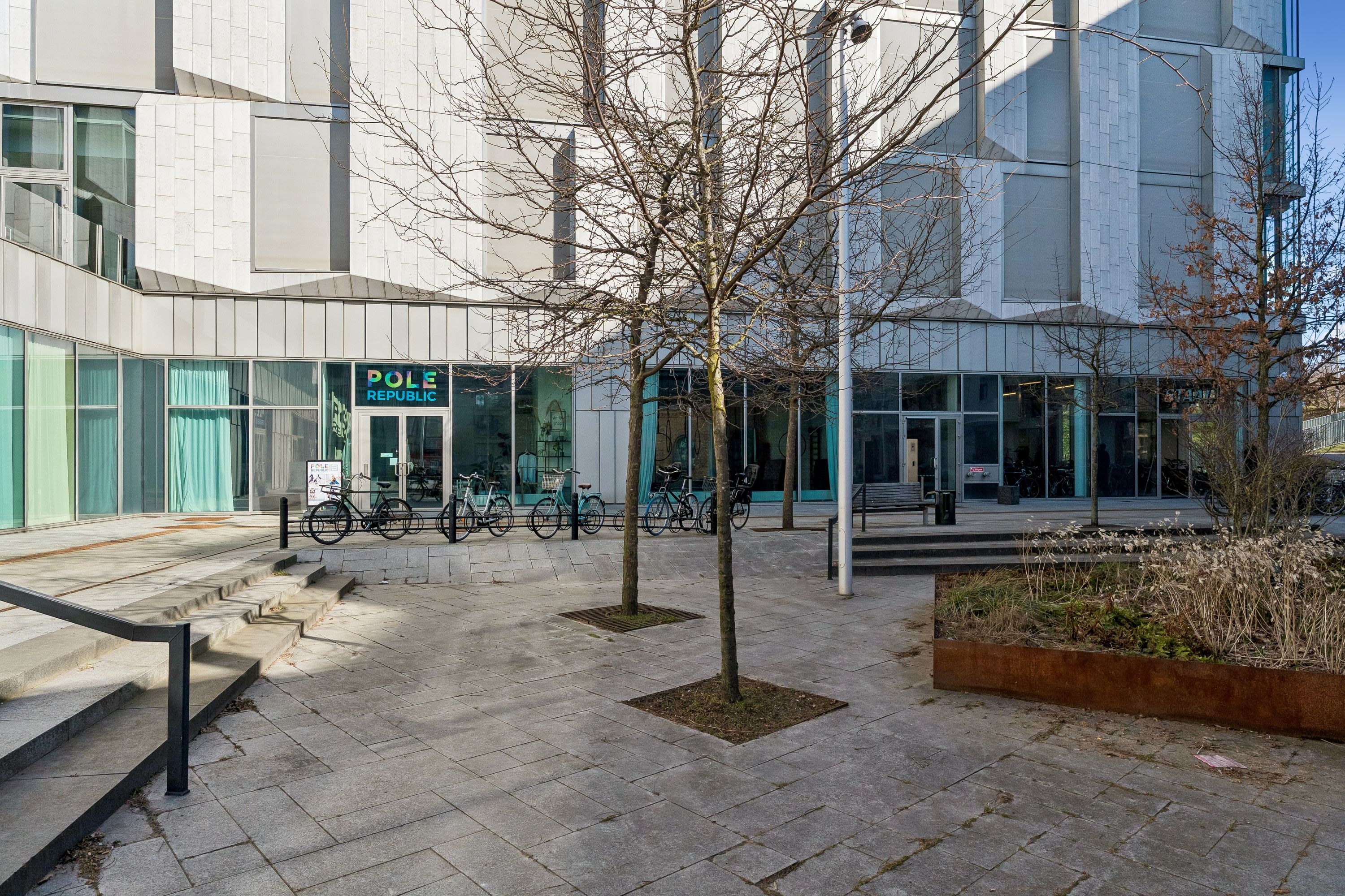 Forretningslokaler med vinduer og indgang til Pole Republic. Parkerede cykler, terrasse, træer og grønne anlæg. Moderne bygning med glasvinduer og mønstret facade