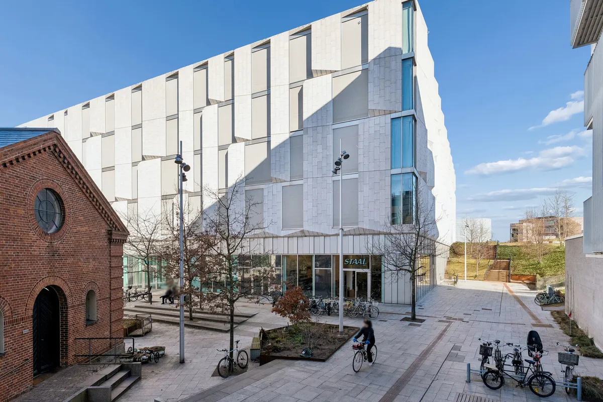 En stor, hvid bygning med store ruder og store, hvide facader og træer uden for. En cykelstativ med fire cykler uden for. En person kører en cykel og parkerer den på cykelstativet.