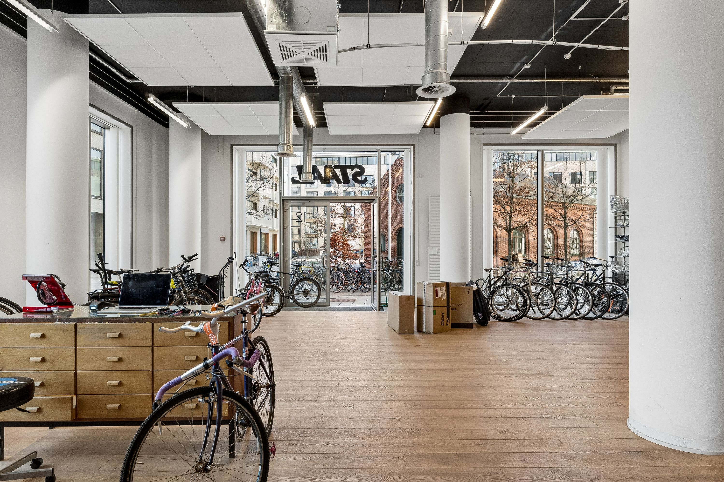 Store, lyse lokaler med moderne indretning, flere cykler stillet op og en skrivebord med laptop. Store vinduer og lyse loftsområder.