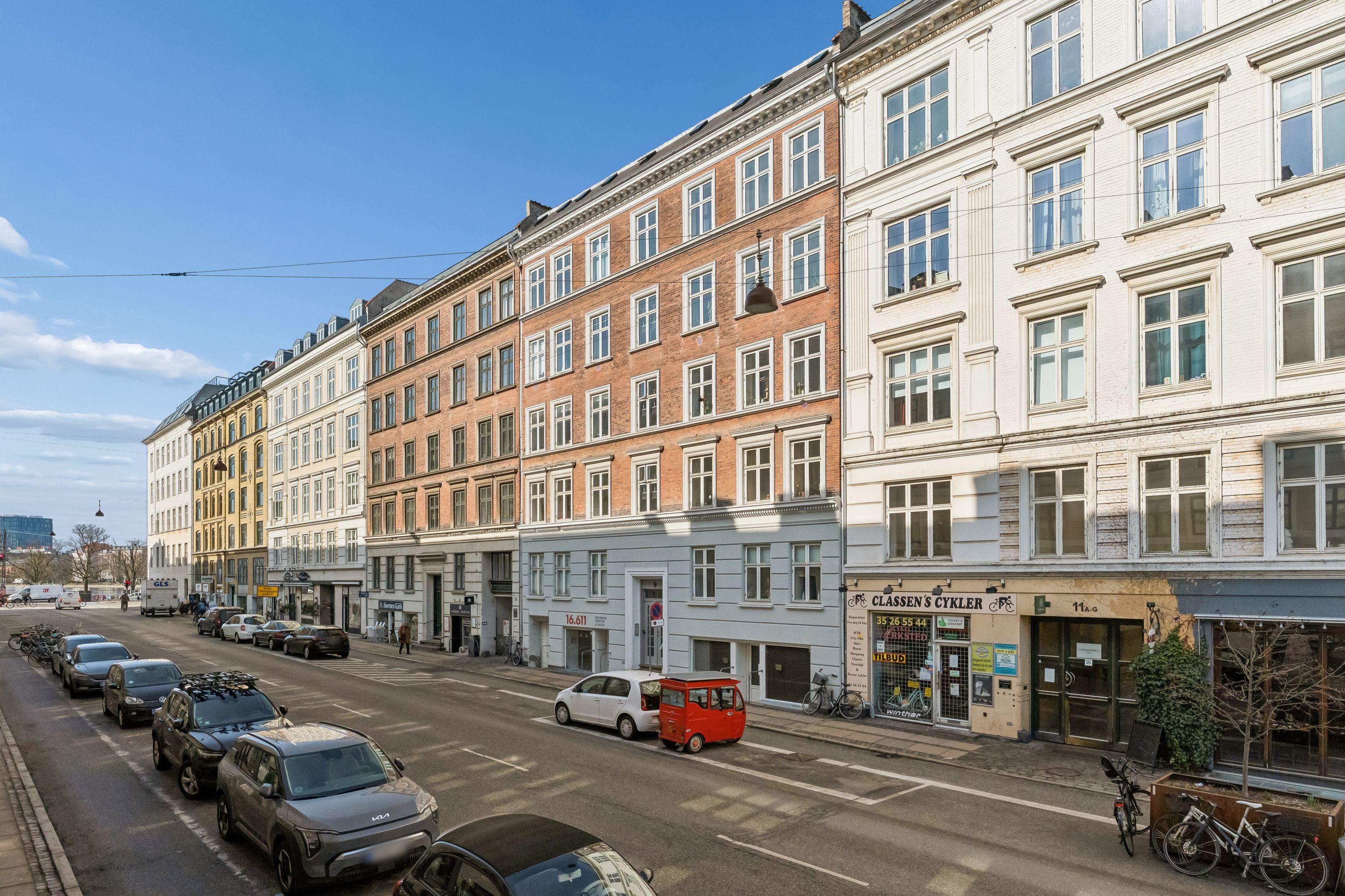 Butikslokaler med store vinduer og røde mursten samt biler parkeret langs gaden og cykler udenfor butikken