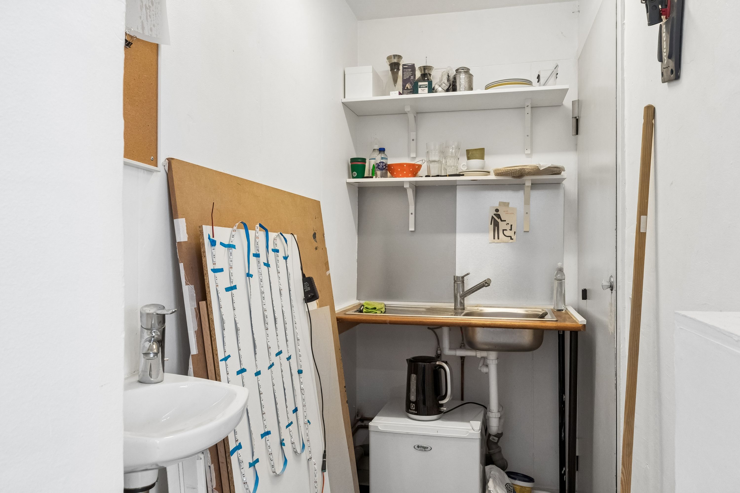 Et lille køkken med hvid væg og gulv, hvor der er et skab og en håndvask med vandhane. Der er et skarpt brædt og et strygebræt hængende på væggen