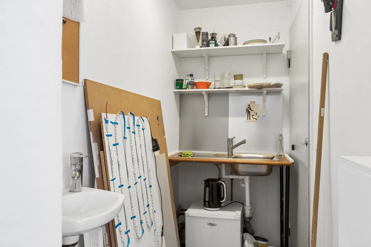 Et lille køkken med hvid væg og gulv, hvor der er et skab og en håndvask med vandhane. Der er et skarpt brædt og et strygebræt hængende på væggen