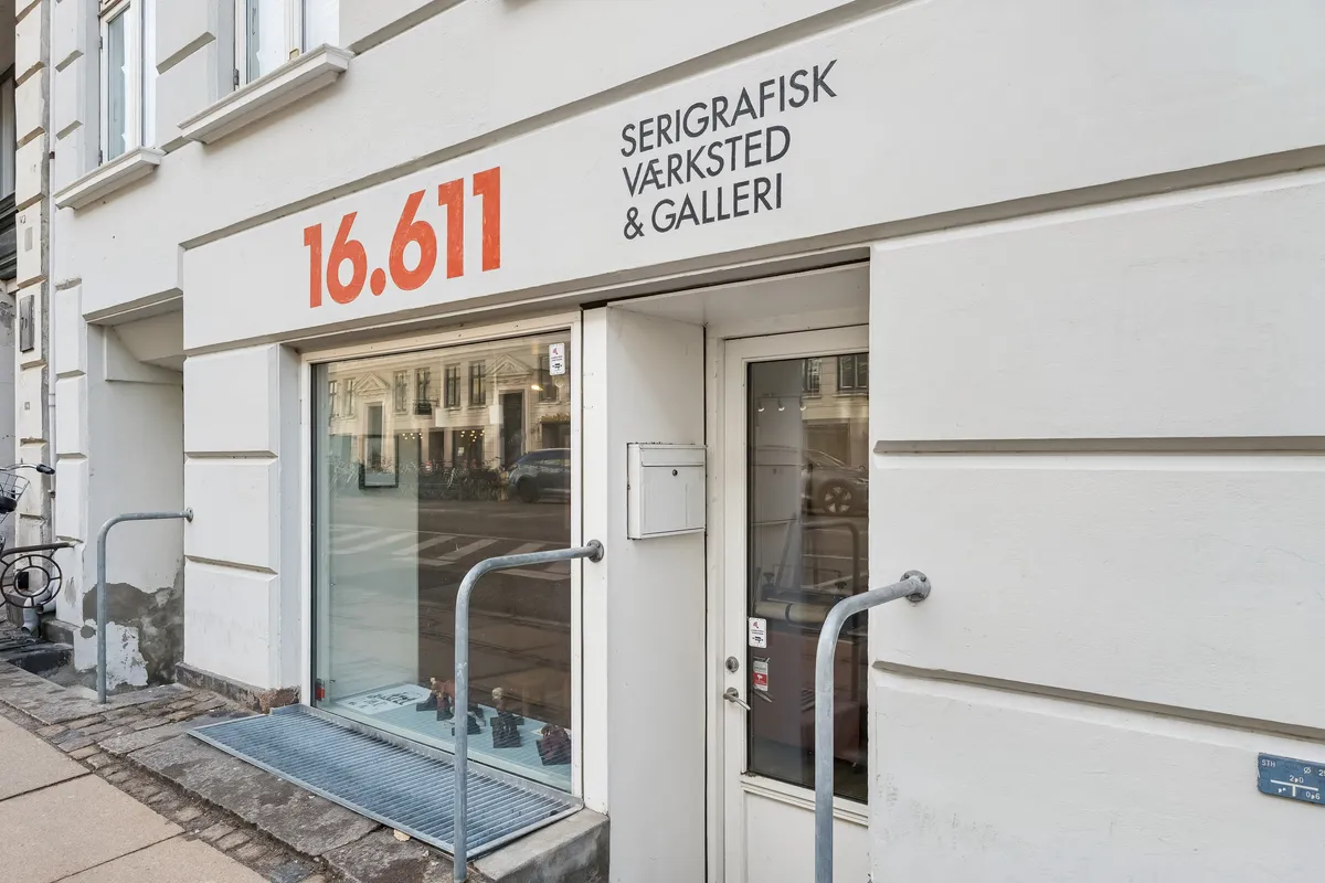 Butiksfront med sort nummer 16.611 og teksten Serigrafisk værksted & galleri. Glassdøre og vinduer. Metalhåndtag og trapper. Vinduesrefleksioner af biler og bygninger