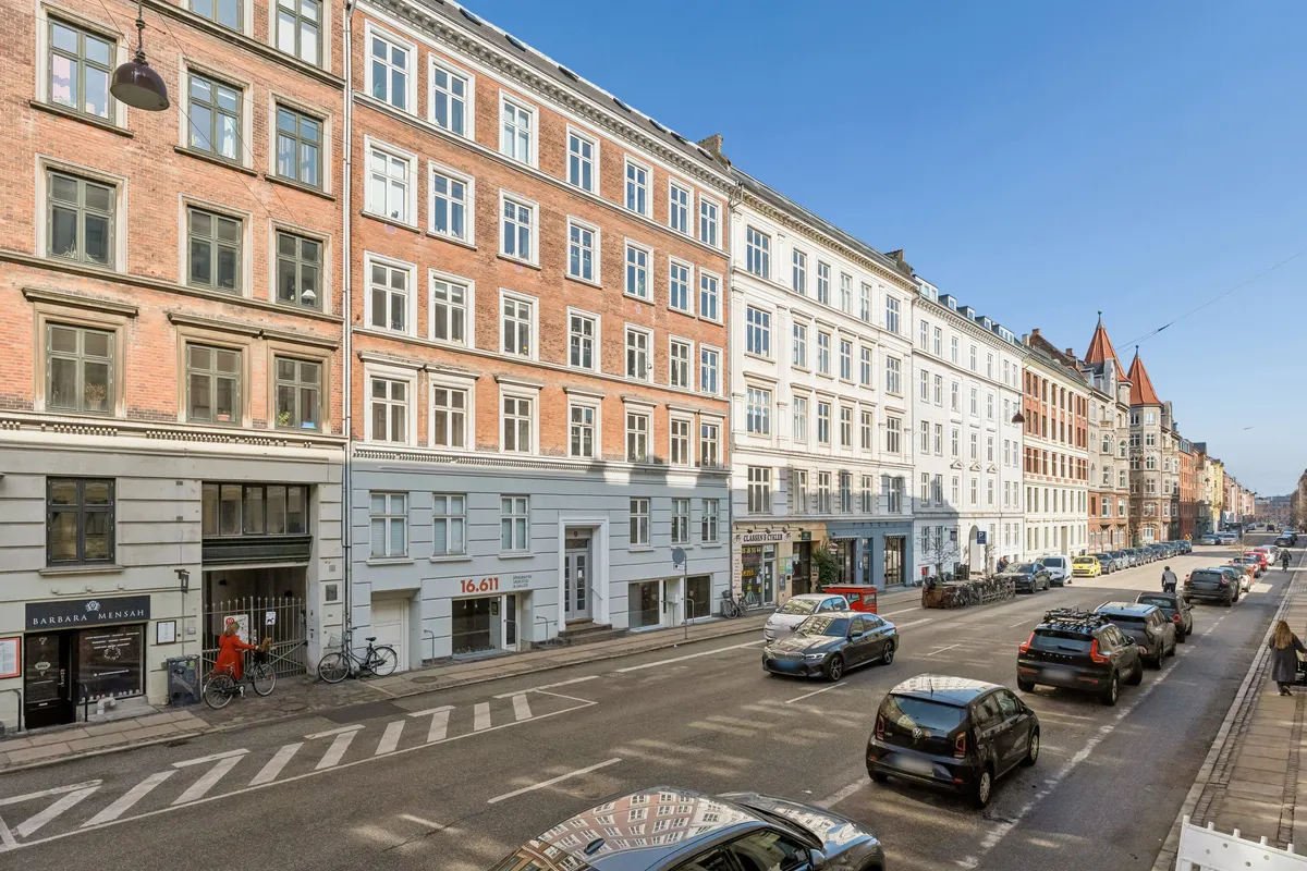 En bilparkering med mange biler og to cykler samt mennesker på gågaden. Bygninger med mange vinduer og en butik med en sort skilt