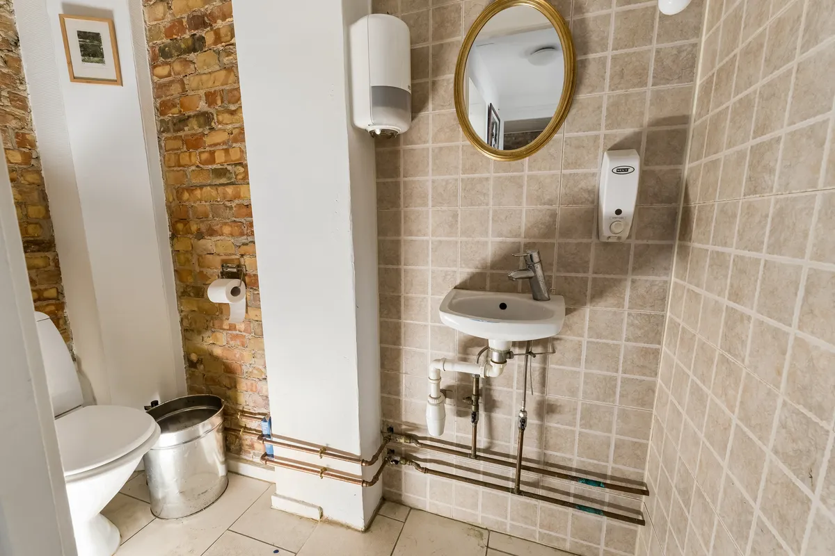 Billet på toilettet med toiletpapir, et håndvask med sæbeautomat og spejl. Toilet med gulv med rør og rørinstallationer og murstensvæg.