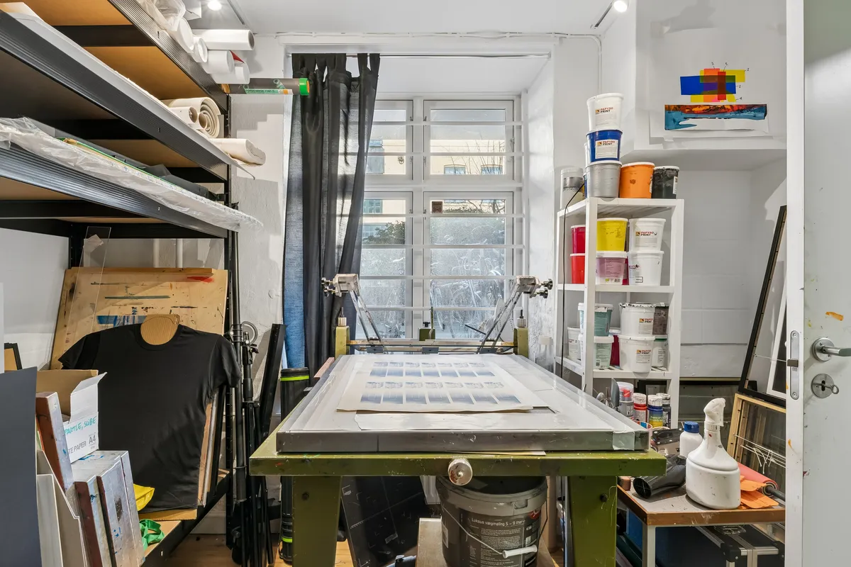 Kunstværkstøjle og materialer i et studie med solbelyst vindue og højloft. Malerier på væggen, vinduesgardiner og værktøj til kunstnere