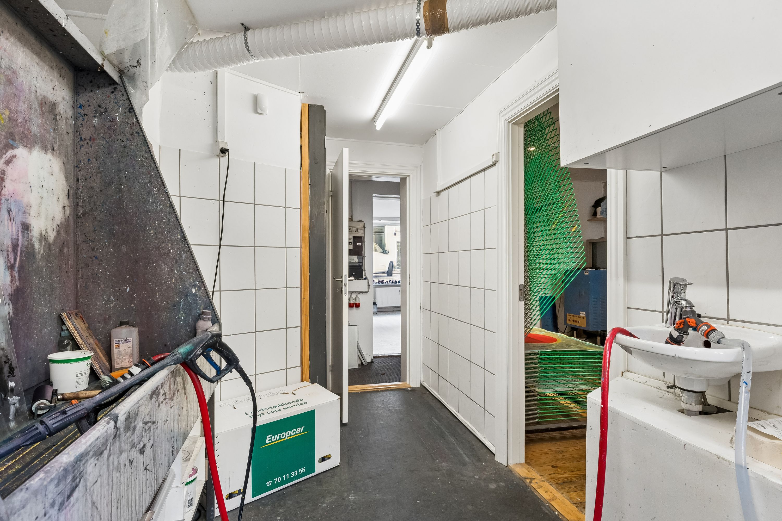 Kort gang med to døre og lyse hvide vægge. Maleri på væggen, stående lysestage og vaskebækken med slange. Diverse materialer og værktøj som børster og rulle.