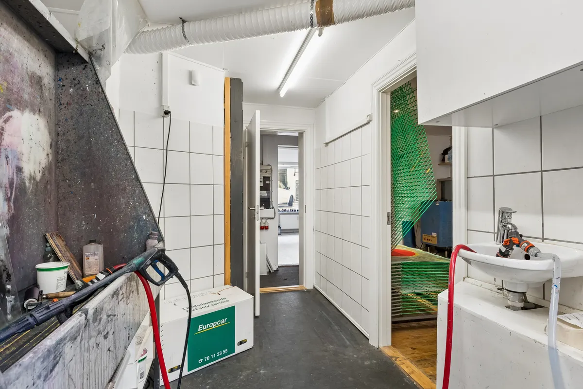 Kort gang med to døre og lyse hvide vægge. Maleri på væggen, stående lysestage og vaskebækken med slange. Diverse materialer og værktøj som børster og rulle.