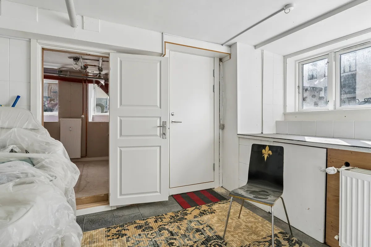 Indvendigt i et rum med hvide vægge og gulv ses en åben dør og gulvtæppe samt en stol og et radiatorpanel.