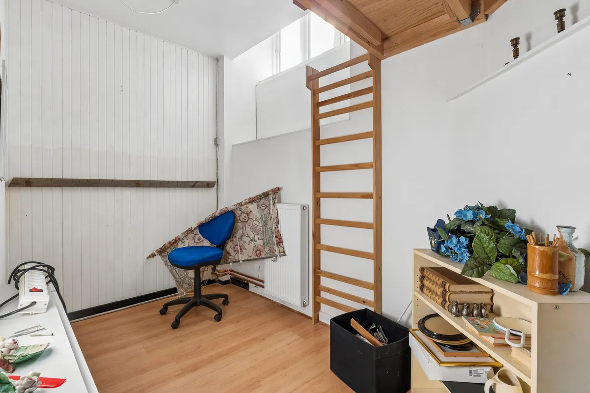 Billede af et værksted med træbrædder, højloft, stol, plante, bøger, smykker, skål, skraldespand og højreol med diverse genstande