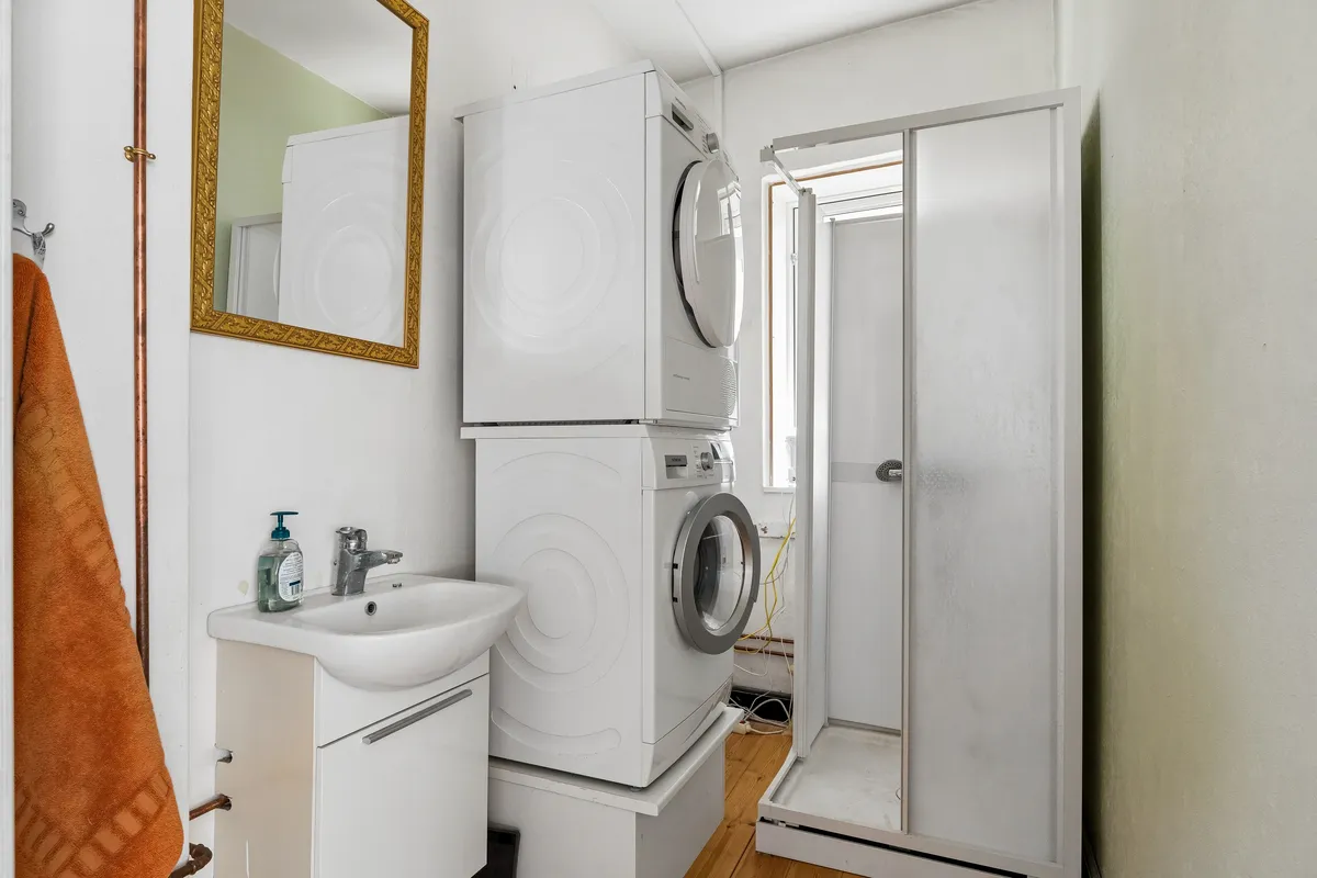 En moderne hvid vaskeri med tørretumbler og vaskemaskine samt en hvid vask og et badeværelse med bruser og spejl