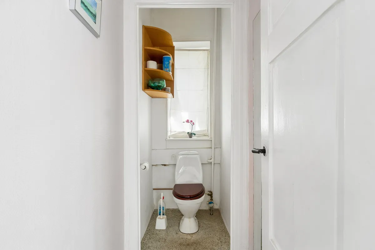 Et lille badeværelse med toilet og toiletpapir, der er ophængt på væggen og smykker på hylden, samt en plante i vinduet