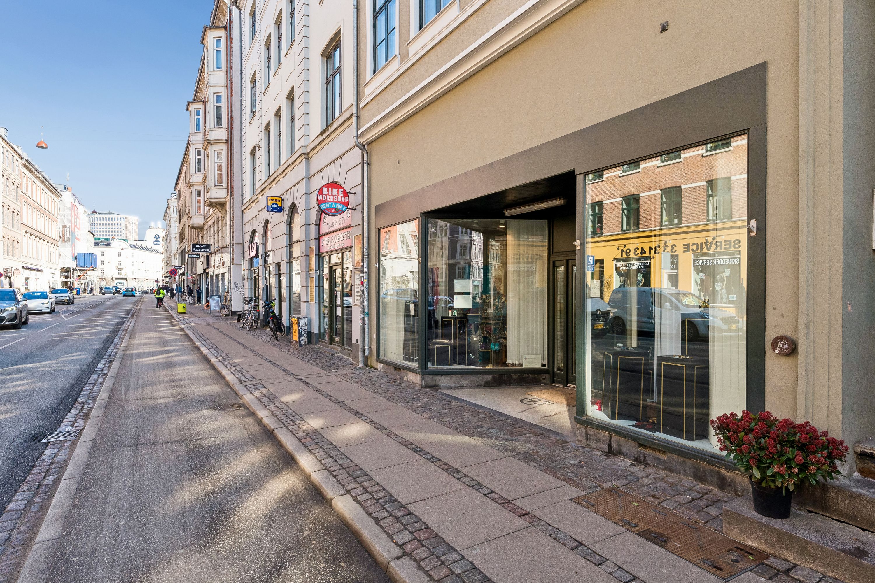 En tom handelsgade med biler og mennesker. En butik med røde blomster, der sælger smykker og ur. Et reklameskilt for en cykelforretning. En høj bygning med mange vinduer.