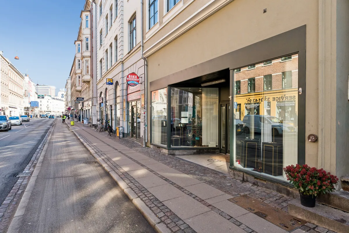En tom handelsgade med biler og mennesker. En butik med røde blomster, der sælger smykker og ur. Et reklameskilt for en cykelforretning. En høj bygning med mange vinduer.