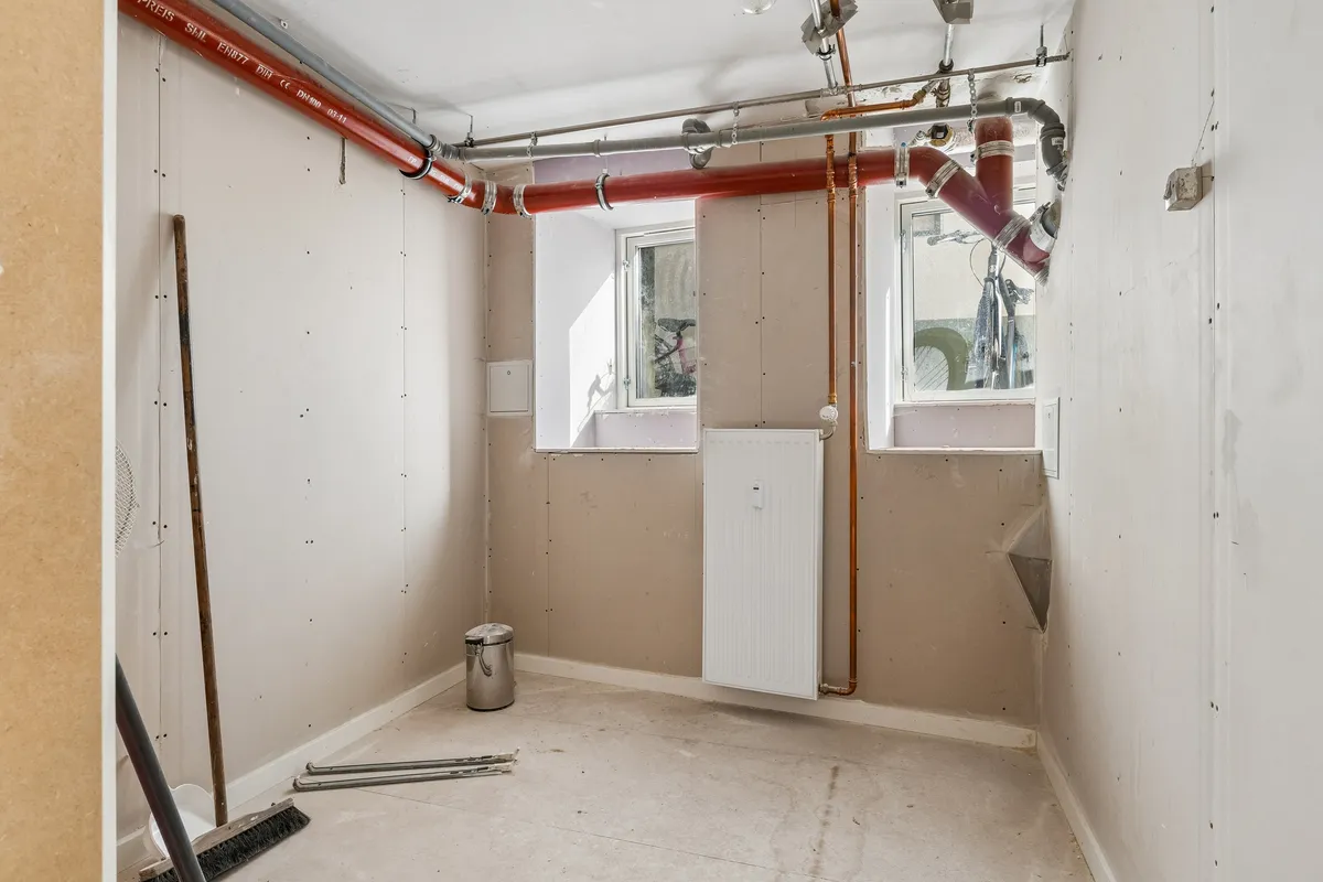 Rum med hvide vægge og rørinstallation samt skraldespand og rengøringsudstyr på gulvet