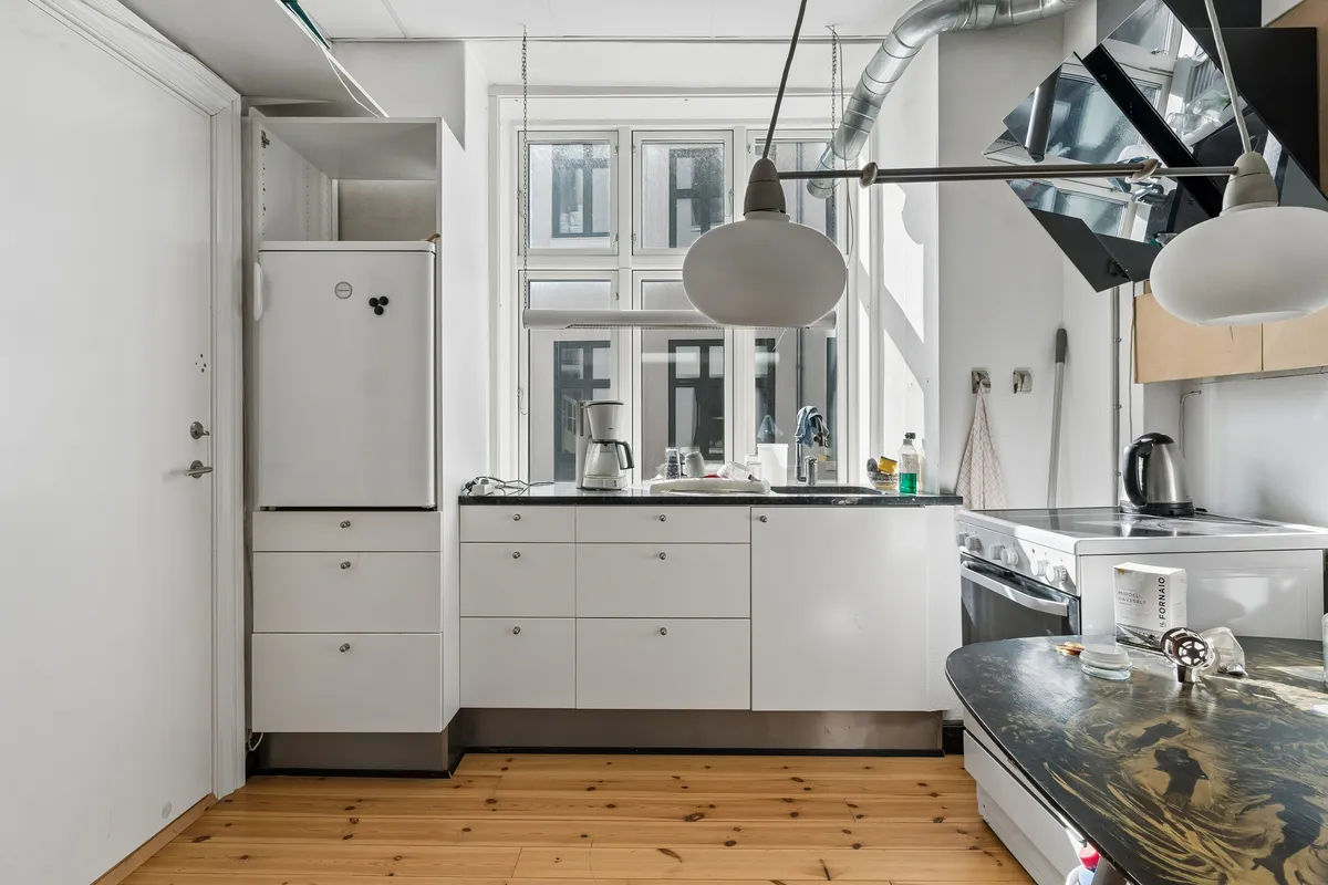 En lille køkken med hvide vægge og gulv, hvidt køleskab, keramik på højden og ovn samt en stor træbordplade med lyse og mørke nuancer