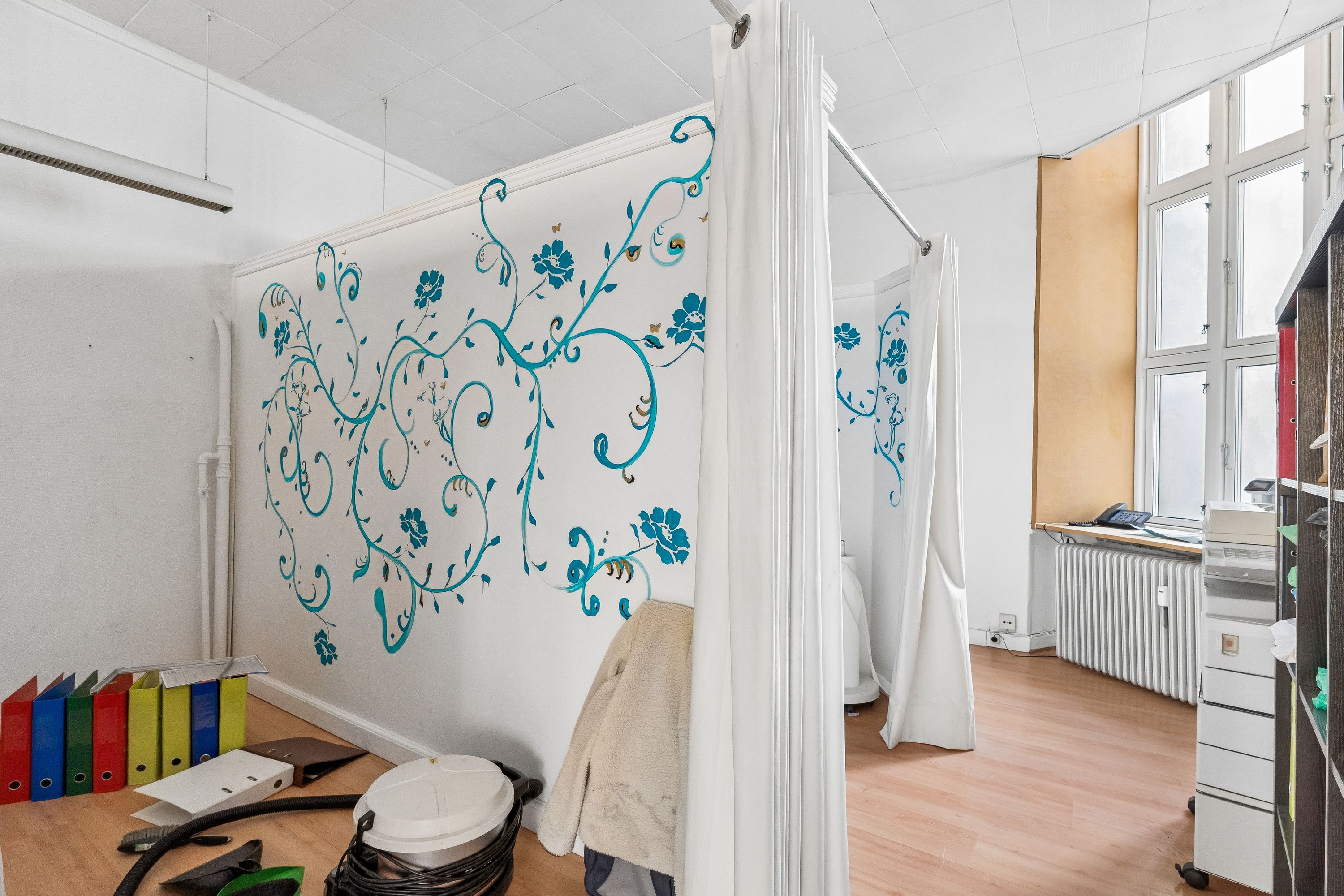 Et lille rum med trægulv, møbler og en hvid væg med blå blomster og ranker. Vinduet og en hvid radiator er synlige i baggrunden.