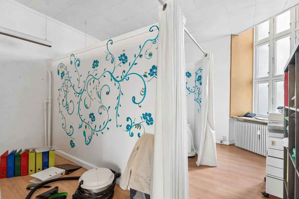 Et lille rum med trægulv, møbler og en hvid væg med blå blomster og ranker. Vinduet og en hvid radiator er synlige i baggrunden.