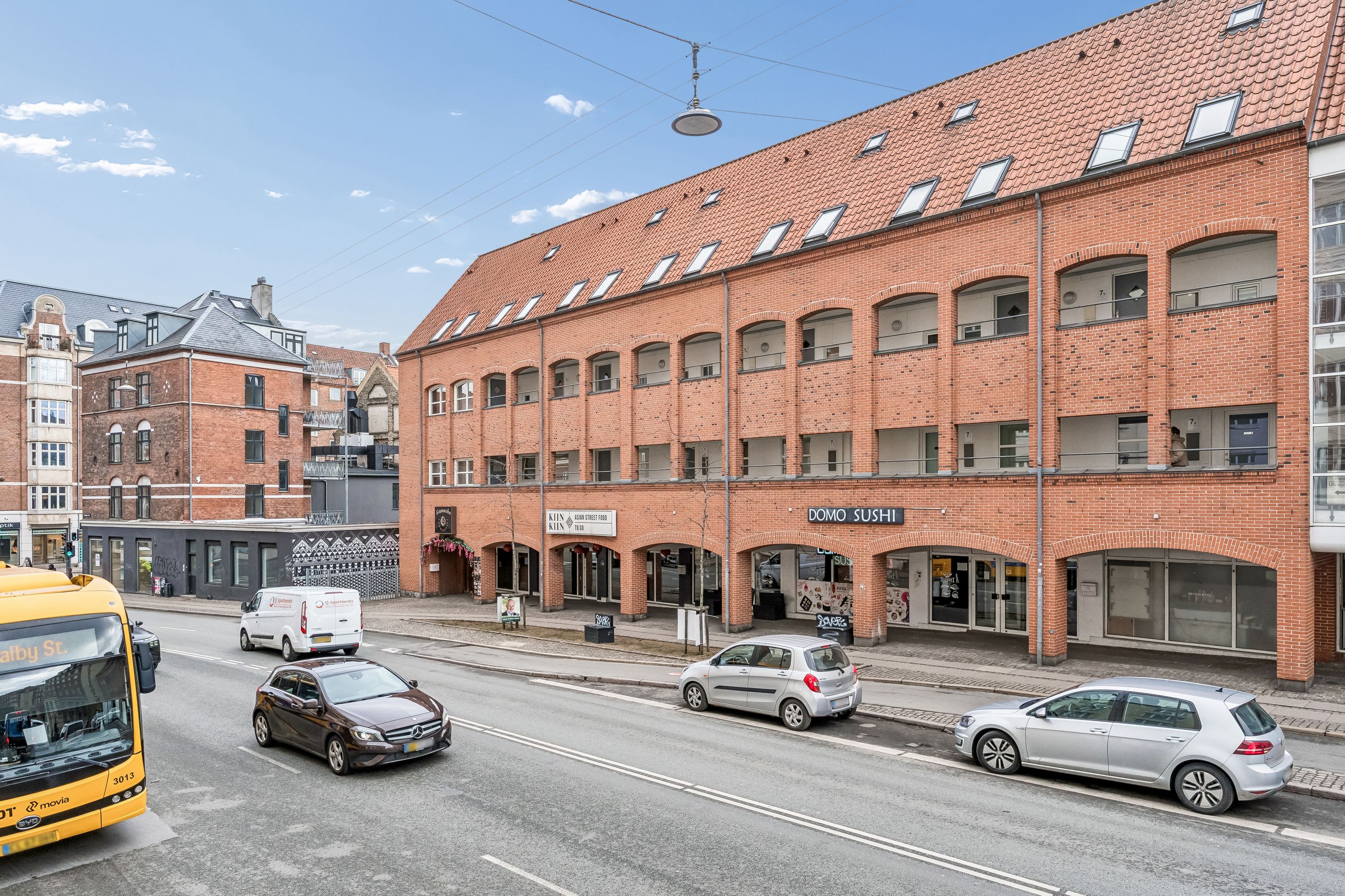 Vejkryds med store biler og bus. Butikslokaler med koreansk mad og sushi i røde mursten. Lys, klar himmel med nogle få skyer.