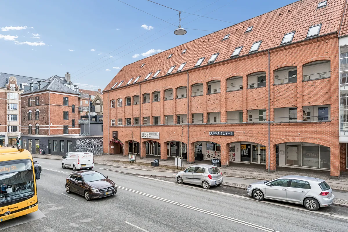 Vejkryds med store biler og bus. Butikslokaler med koreansk mad og sushi i røde mursten. Lys, klar himmel med nogle få skyer.
