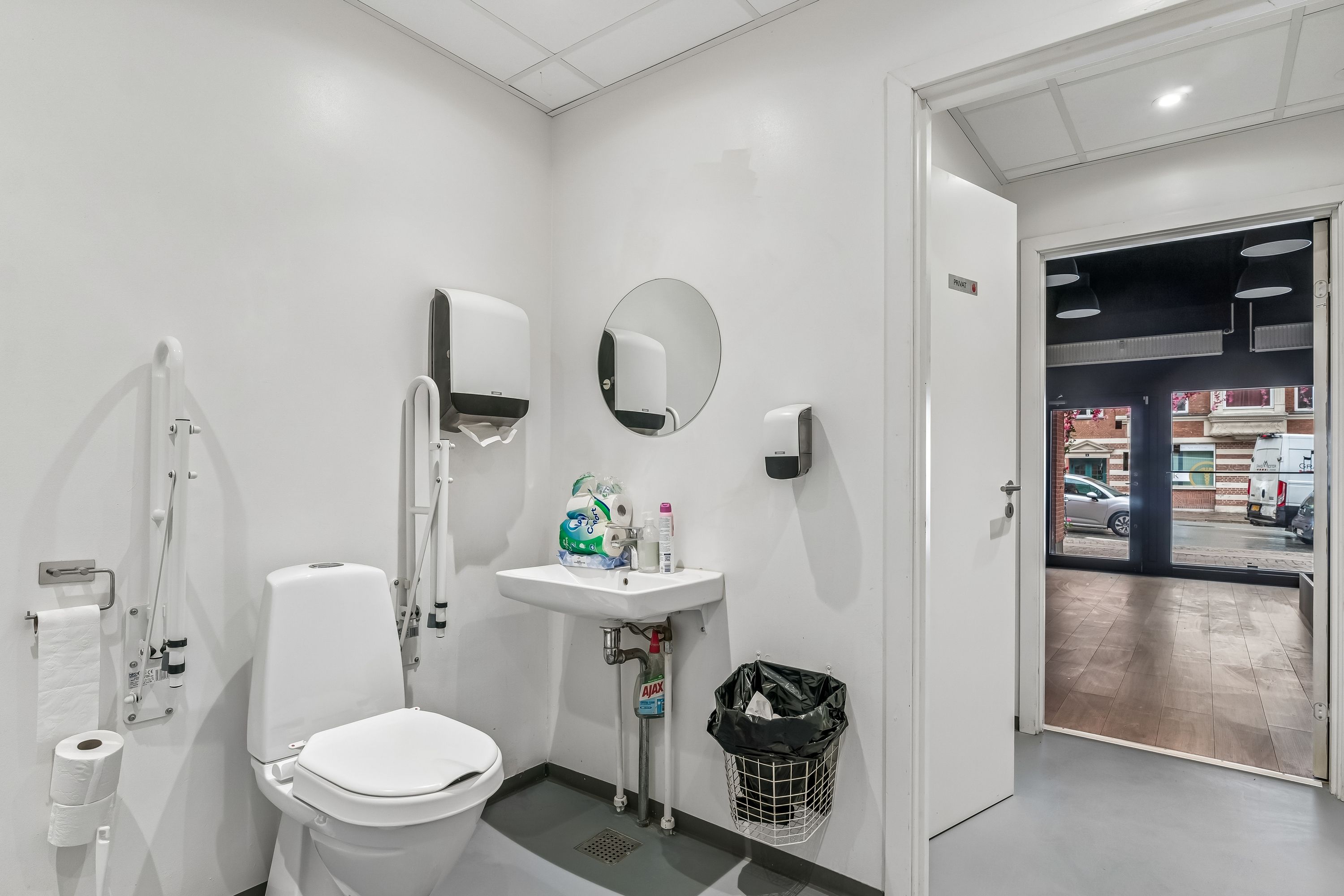 Moderne hvid toiletrum med høj kvalitet. Der er et håndvask med sæbe, et toilet og et sædeskud. Der er et spejl og håndklæder. Der er et spild og en sække.