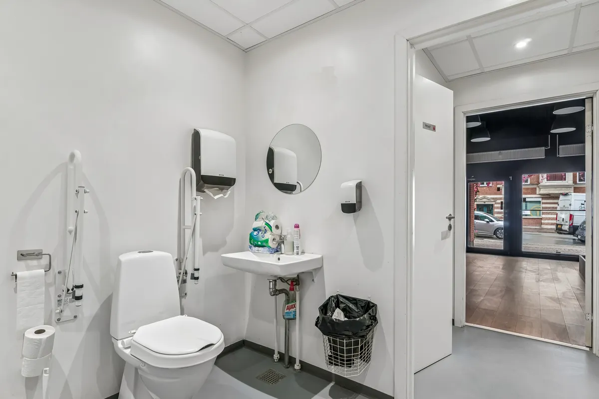 Moderne hvid toiletrum med høj kvalitet. Der er et håndvask med sæbe, et toilet og et sædeskud. Der er et spejl og håndklæder. Der er et spild og en sække.