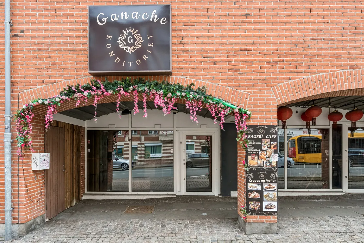En rødmalet murstensbygning med glasdøre og vinduer og en lyserød bue med blomster og en stolpe med en menuliste og en sølvbus i baggrunden