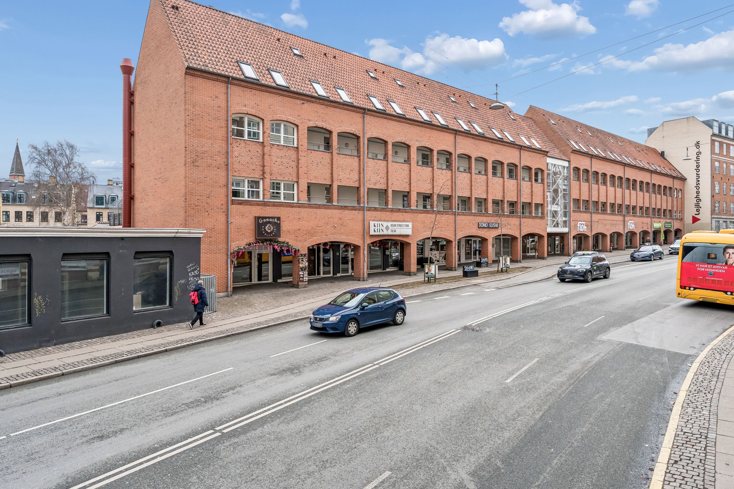 En stor rød murstensbygning med forretninger og et busstop på Toftegårds Allé. En person med rygsæk går langs fortovskanten. Adkomstvej med biler og en tom busstop.